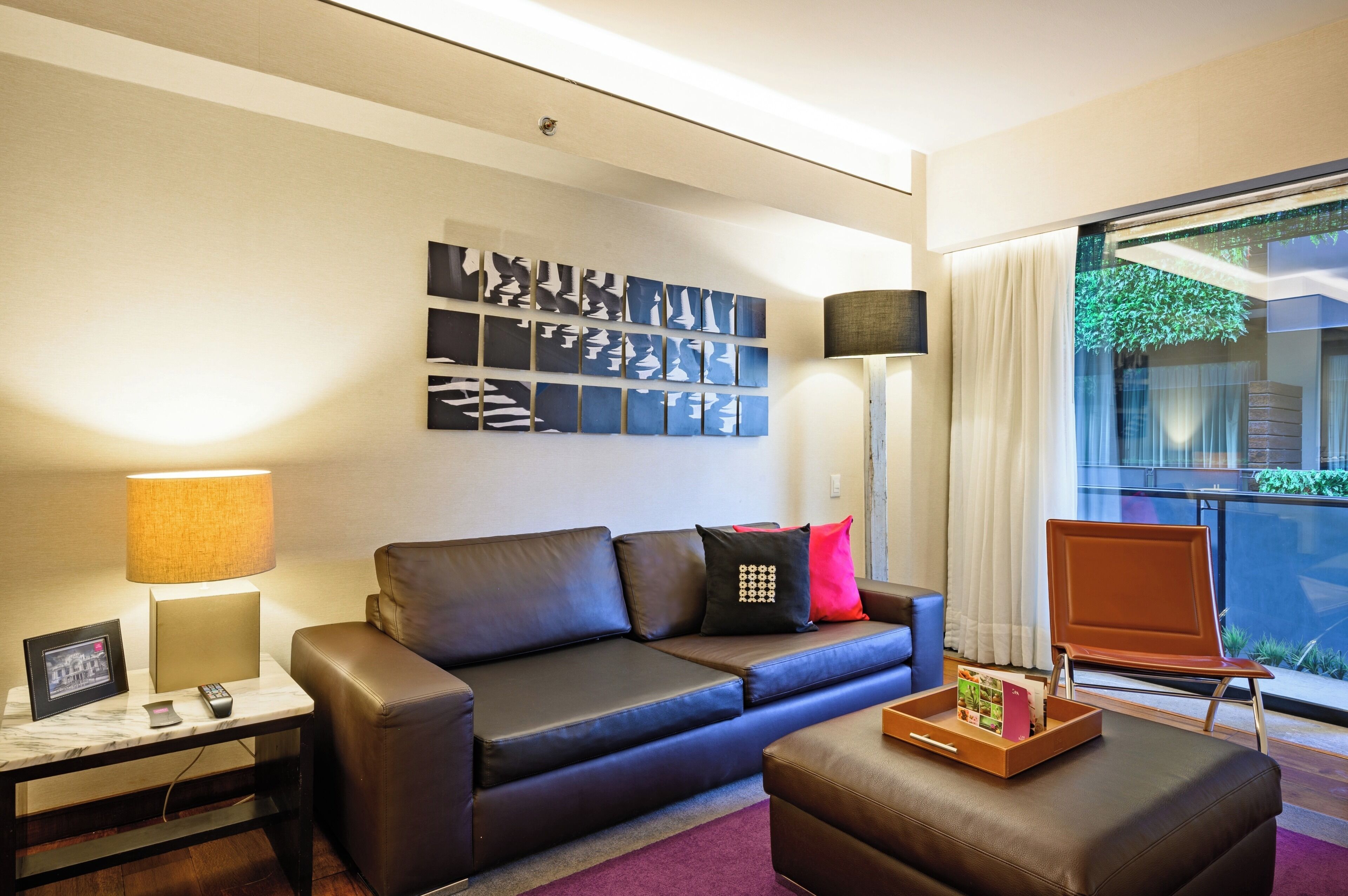 A modern living room with a leather sofa, a side table with a lamp, a tray with books, and a large window with a view of greenery.