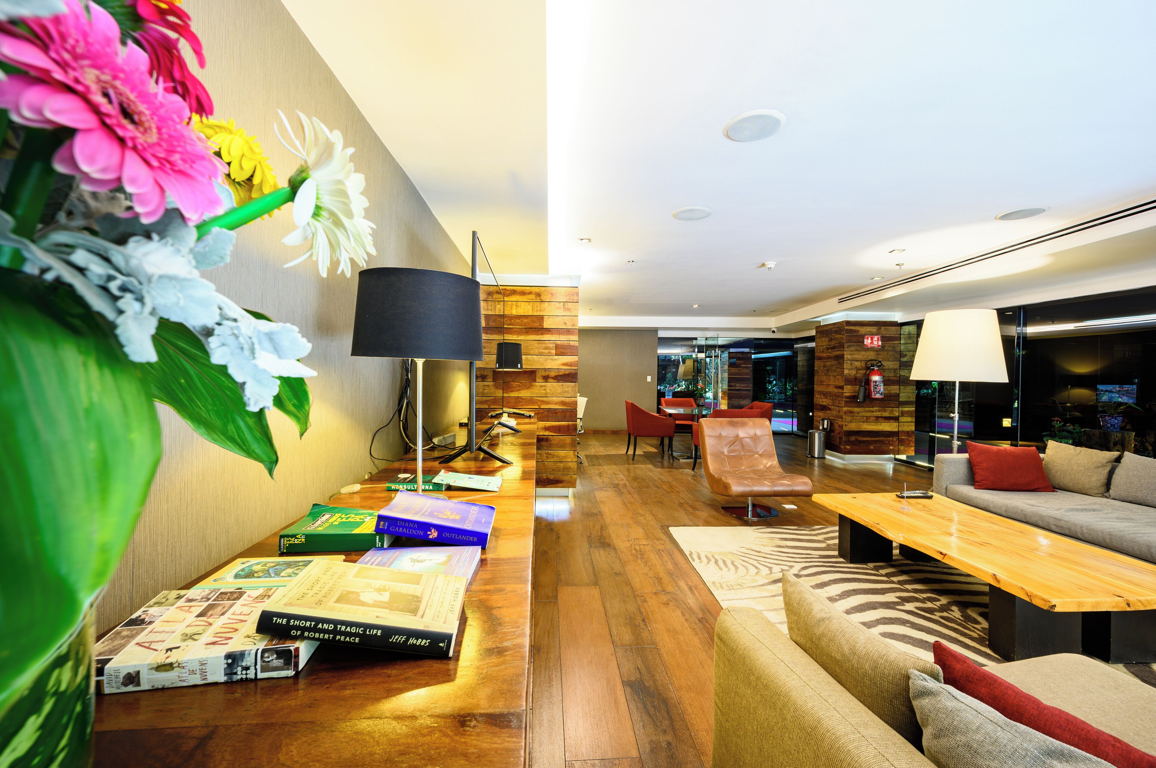 A modern hotel lobby with a wooden reception desk, a sofa, a dining area, and a large floral arrangement.