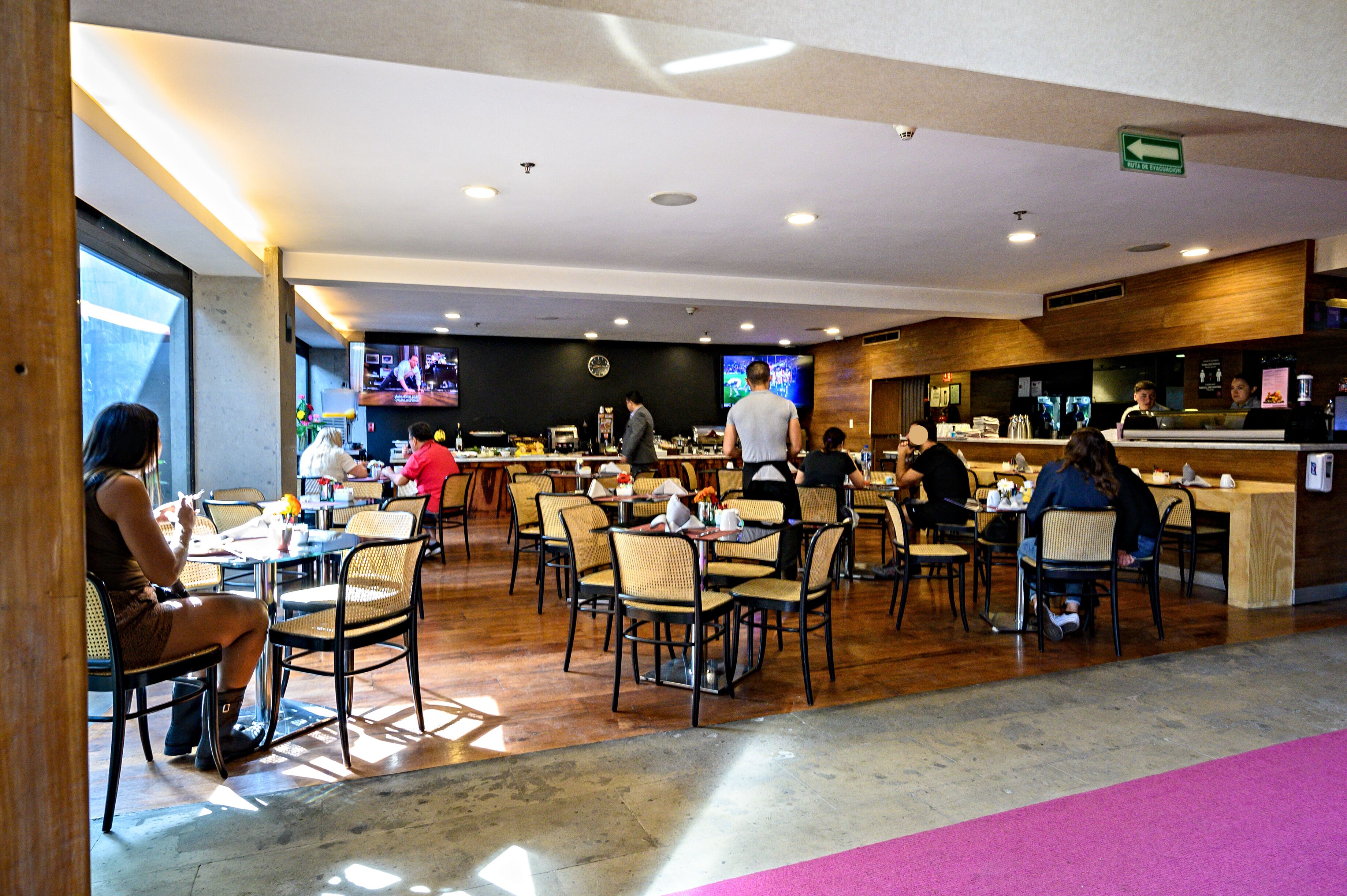 Een ruim restaurant met meerdere tafels en stoelen, een bar en een televisiescherm waarop een programma wordt vertoond.