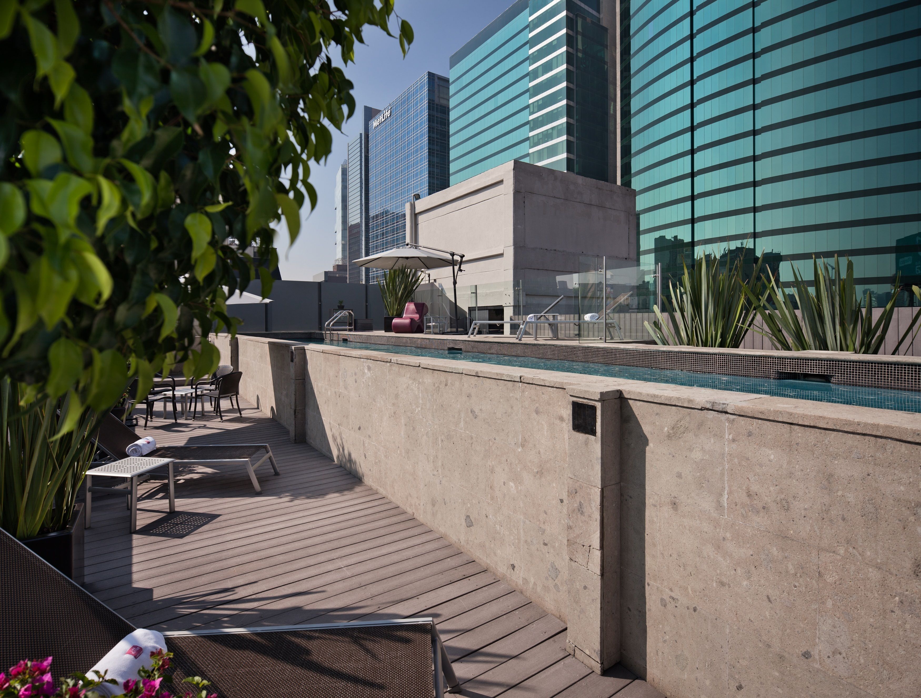 Zona de piscina en la azotea con sillones y una mesa, rodeada de edificios modernos.