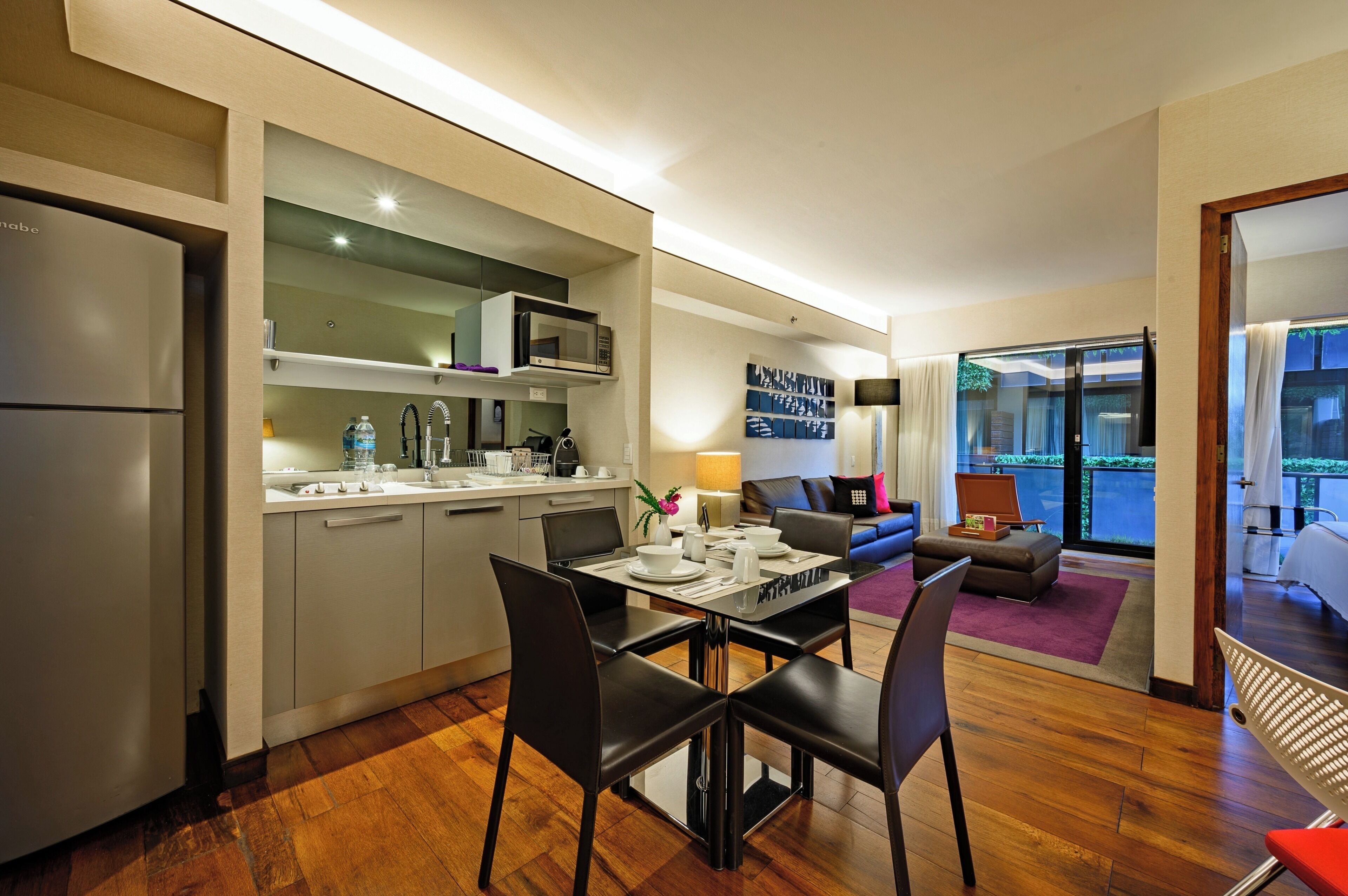 A modern kitchen with a built-in microwave, a dining area with black chairs, and a living room with a sofa and a large window.