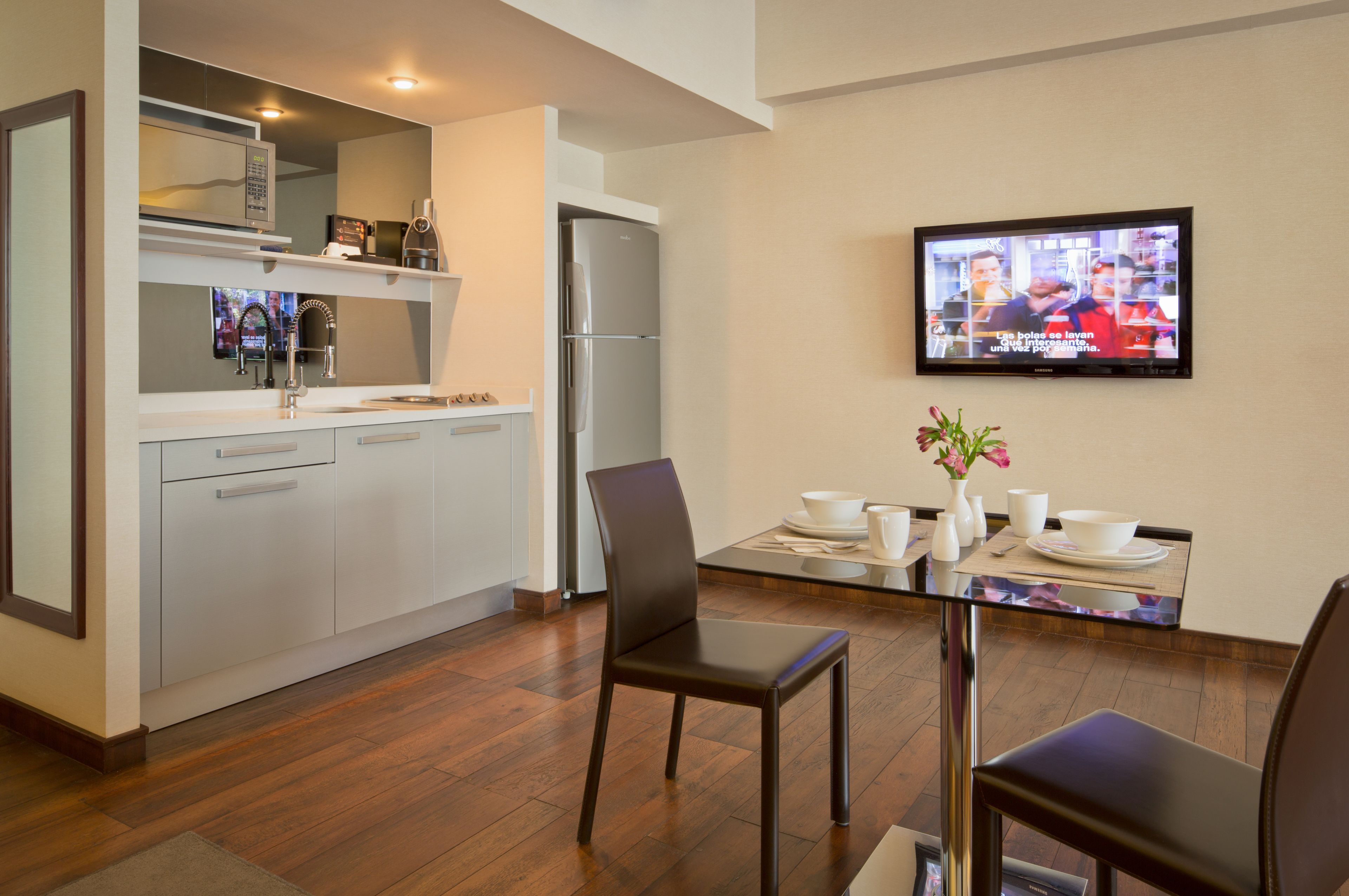 Een moderne keuken met een koelkast, magnetron en gootsteen. Er is een eethoek met een tafel, stoelen en een aan de muur bevestigde televisie.