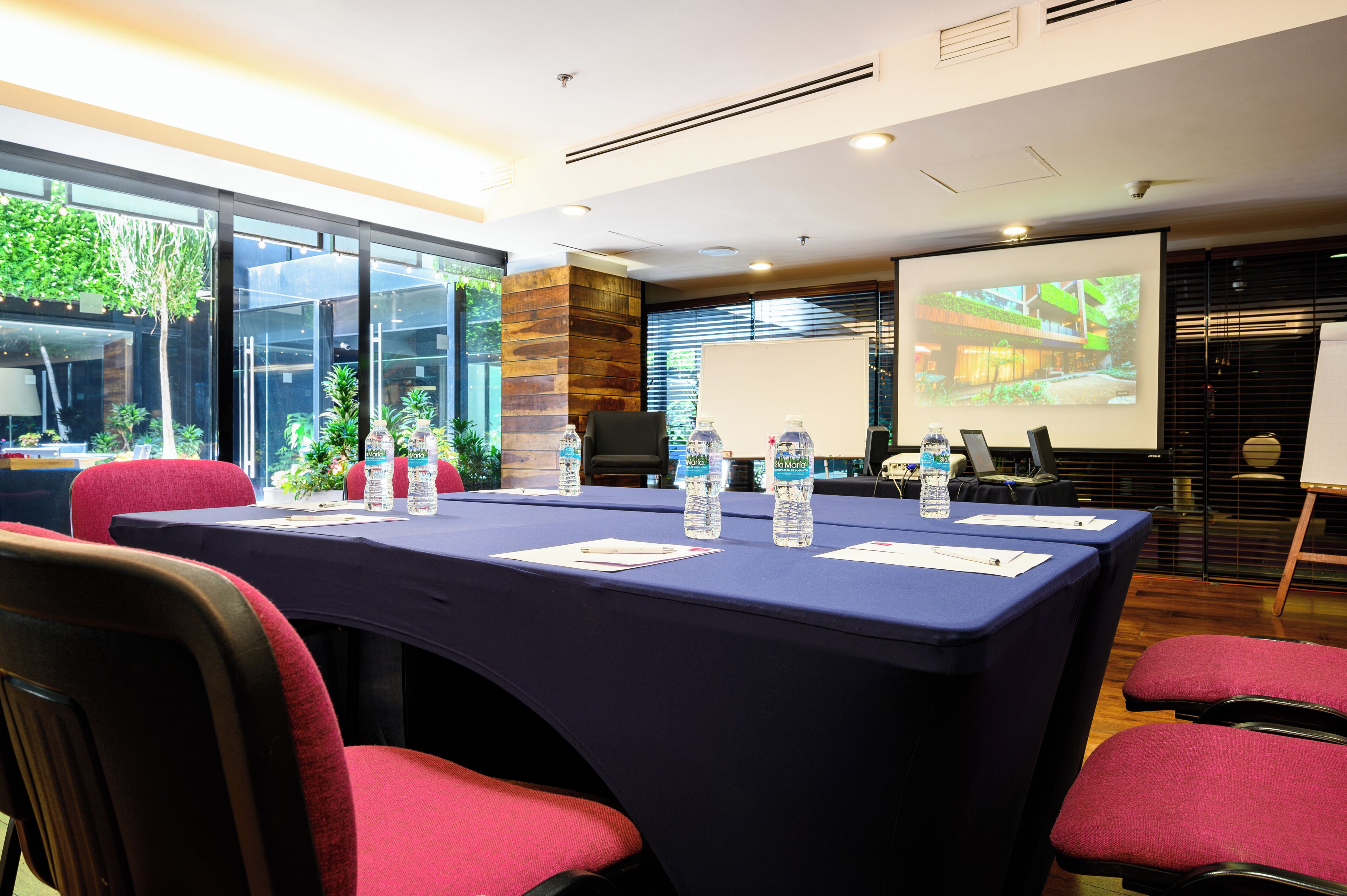 Un salón de conferencias con una gran mesa, sillas, una pantalla de proyector y vistas al exterior con vegetación.