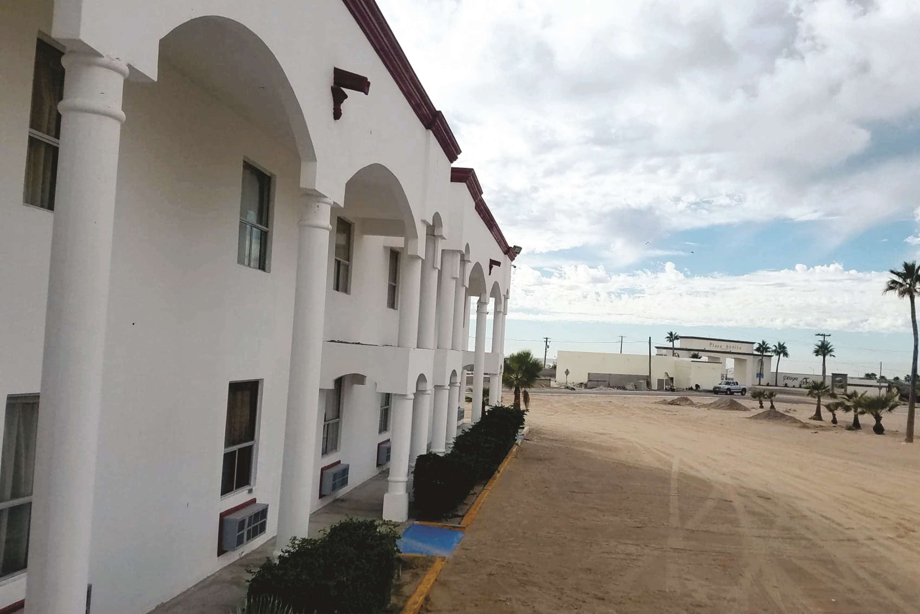 A white building with multiple arches and columns, a sandy ground, and a few palm trees.