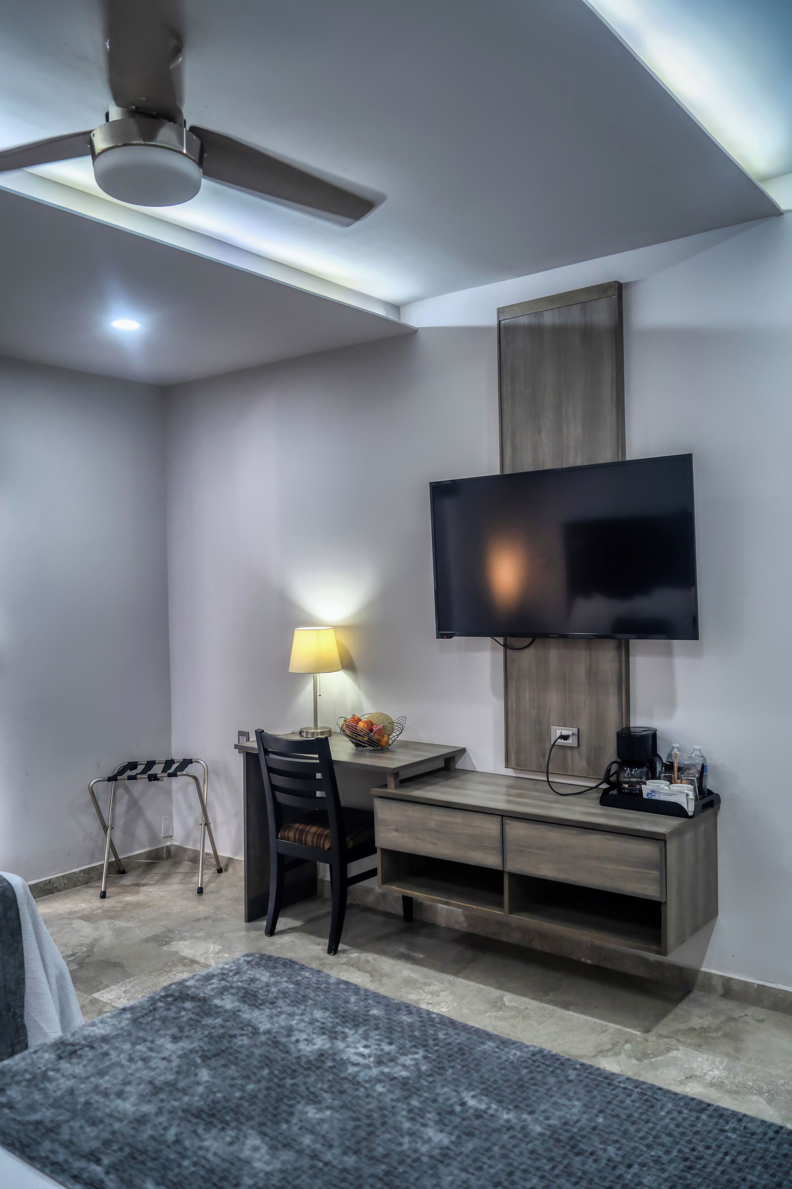 A modern hotel room with a flat-screen TV mounted on the wall, a desk with a lamp, a chair, and a small table with a basket.