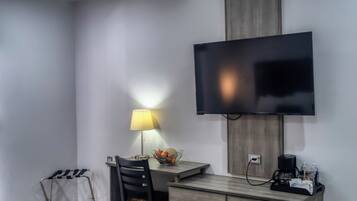 A modern hotel room with a flat-screen TV mounted on the wall, a desk with a lamp, a chair, and a small table with a basket.
