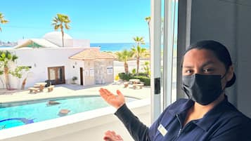 A person standing by a window overlooking a pool area with a view of a building and palm trees.