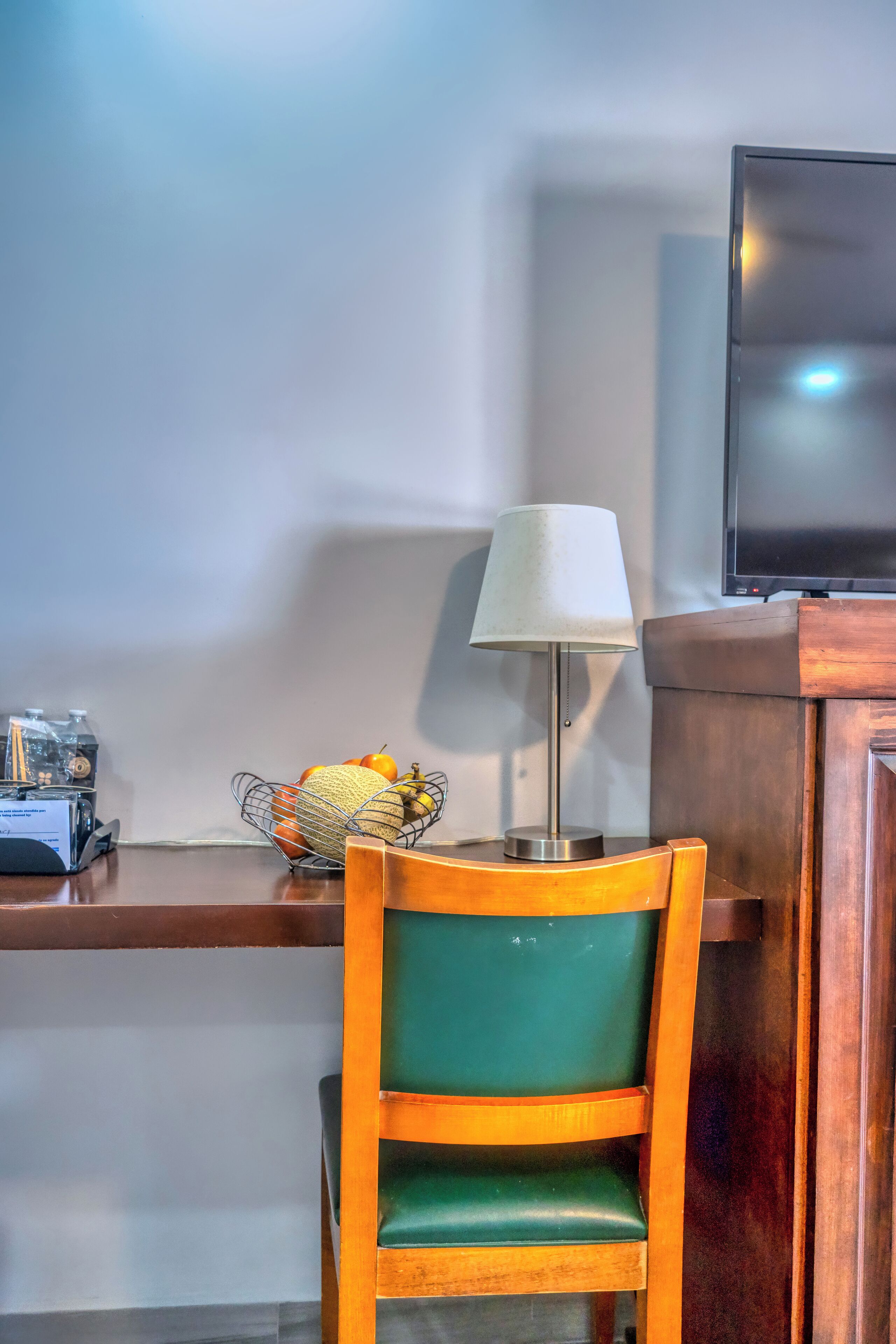 A wooden chair with a green cushion, a metal lamp with a white shade, and a fruit basket on a wooden shelf.