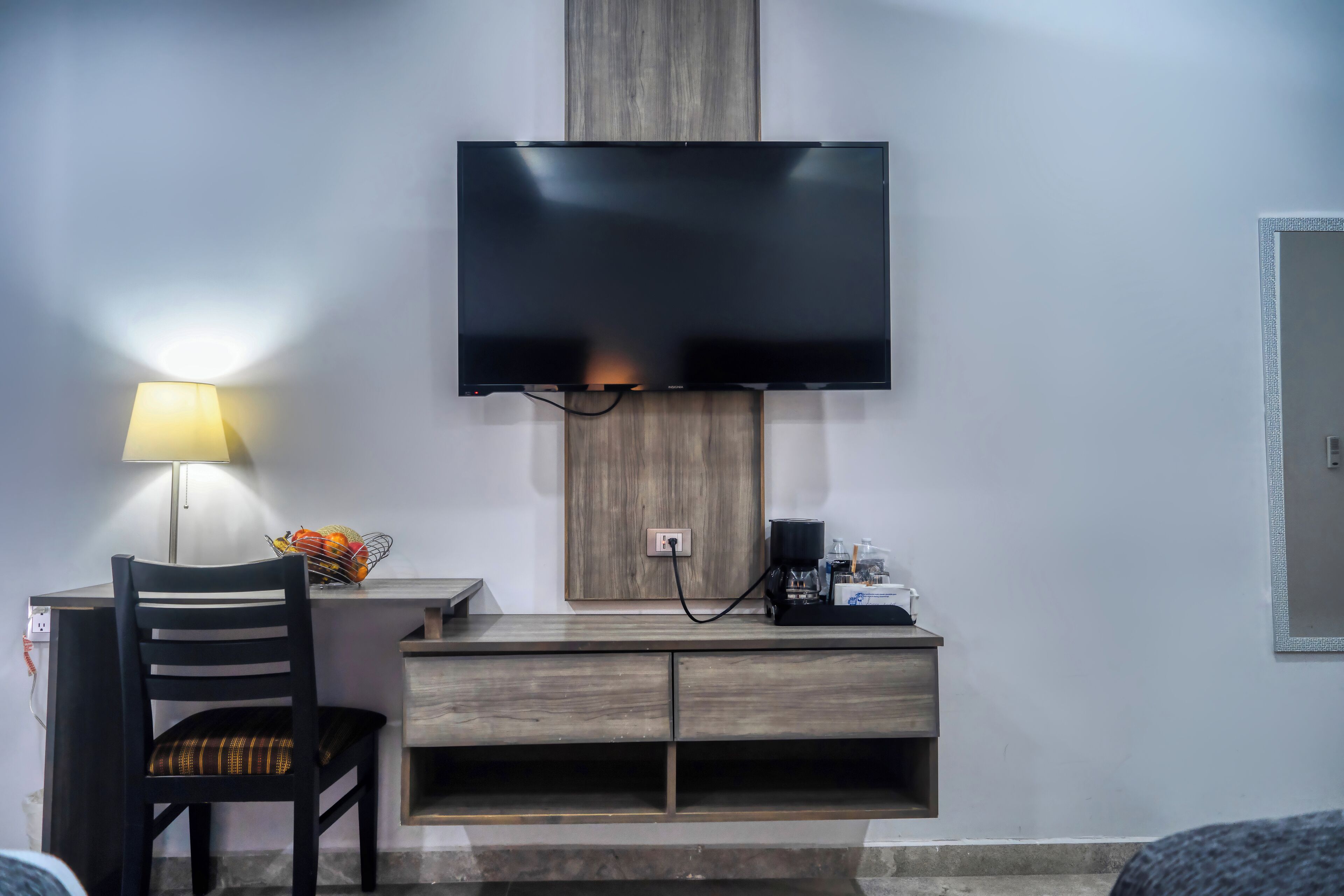 A hotel room with a flat-screen TV mounted on the wall, a desk with a lamp, a chair, and a coffee machine.