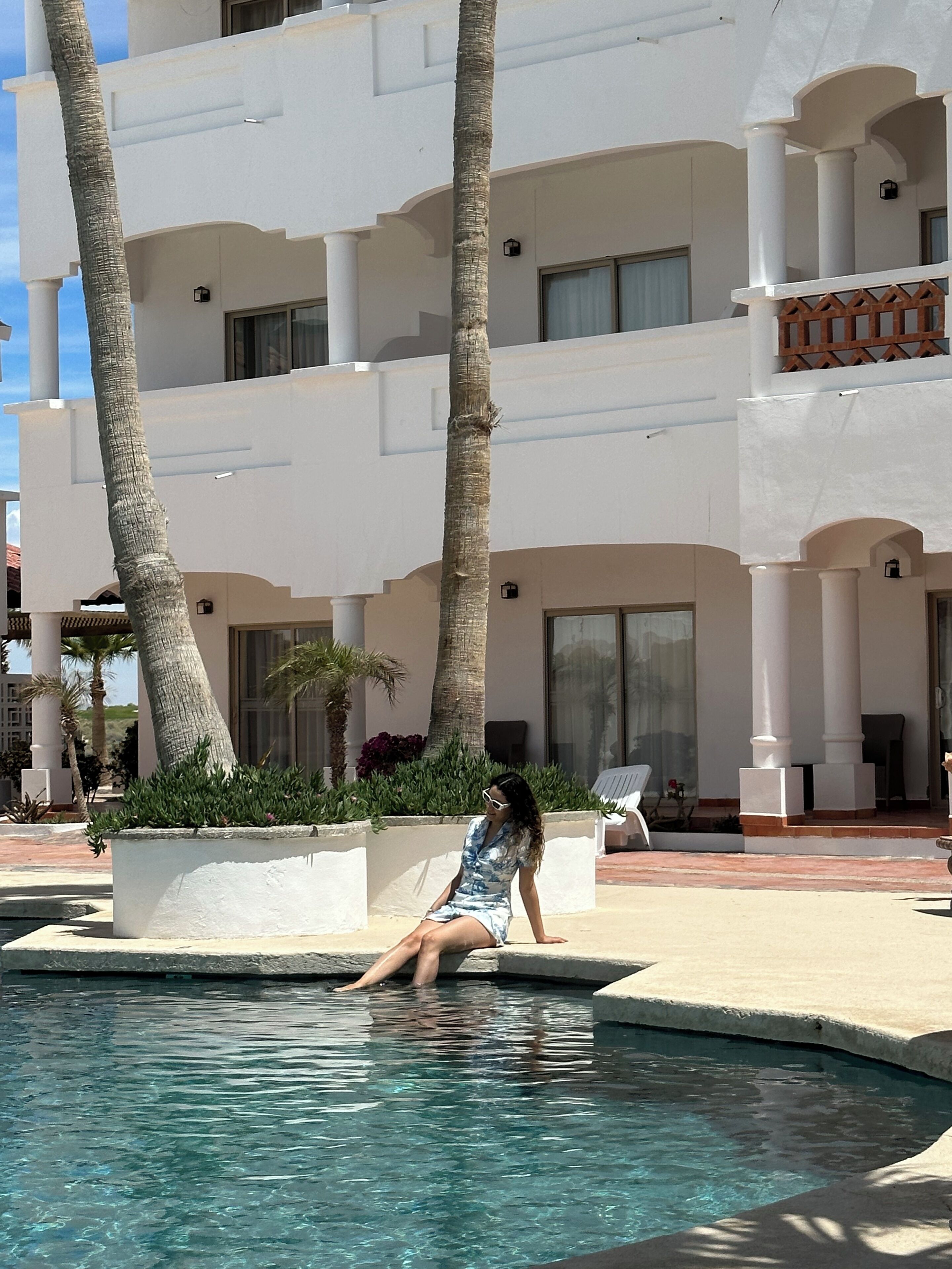 A woman sitting by a pool in a resort setting with white buildings and palm trees.