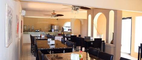 A spacious dining area with multiple tables and chairs, a ceiling fan, and a kitchen in the background.
