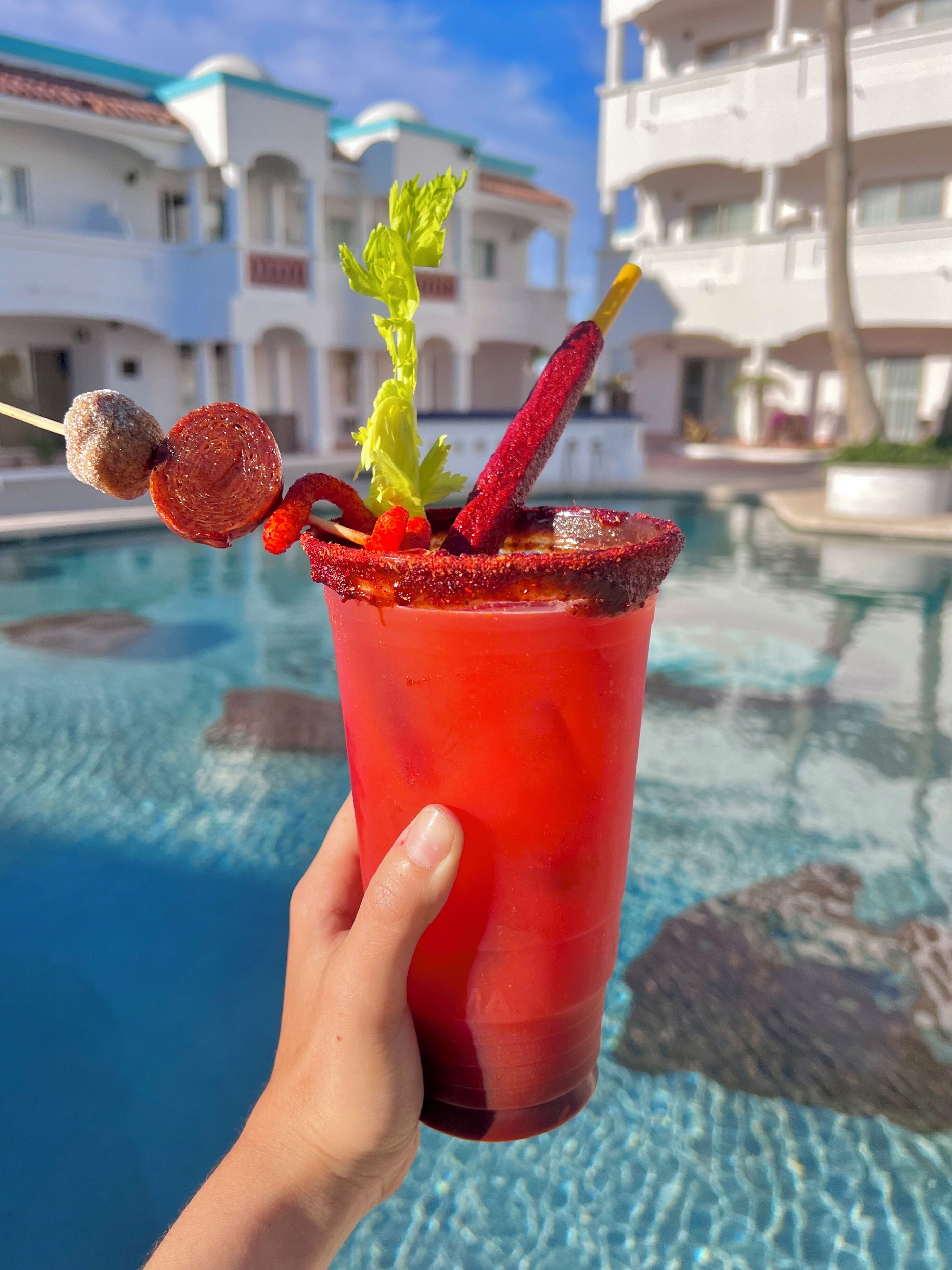 A hand holding a red drink with a garnish, a skewer with a fruit and a candy, and a pool in the background.
