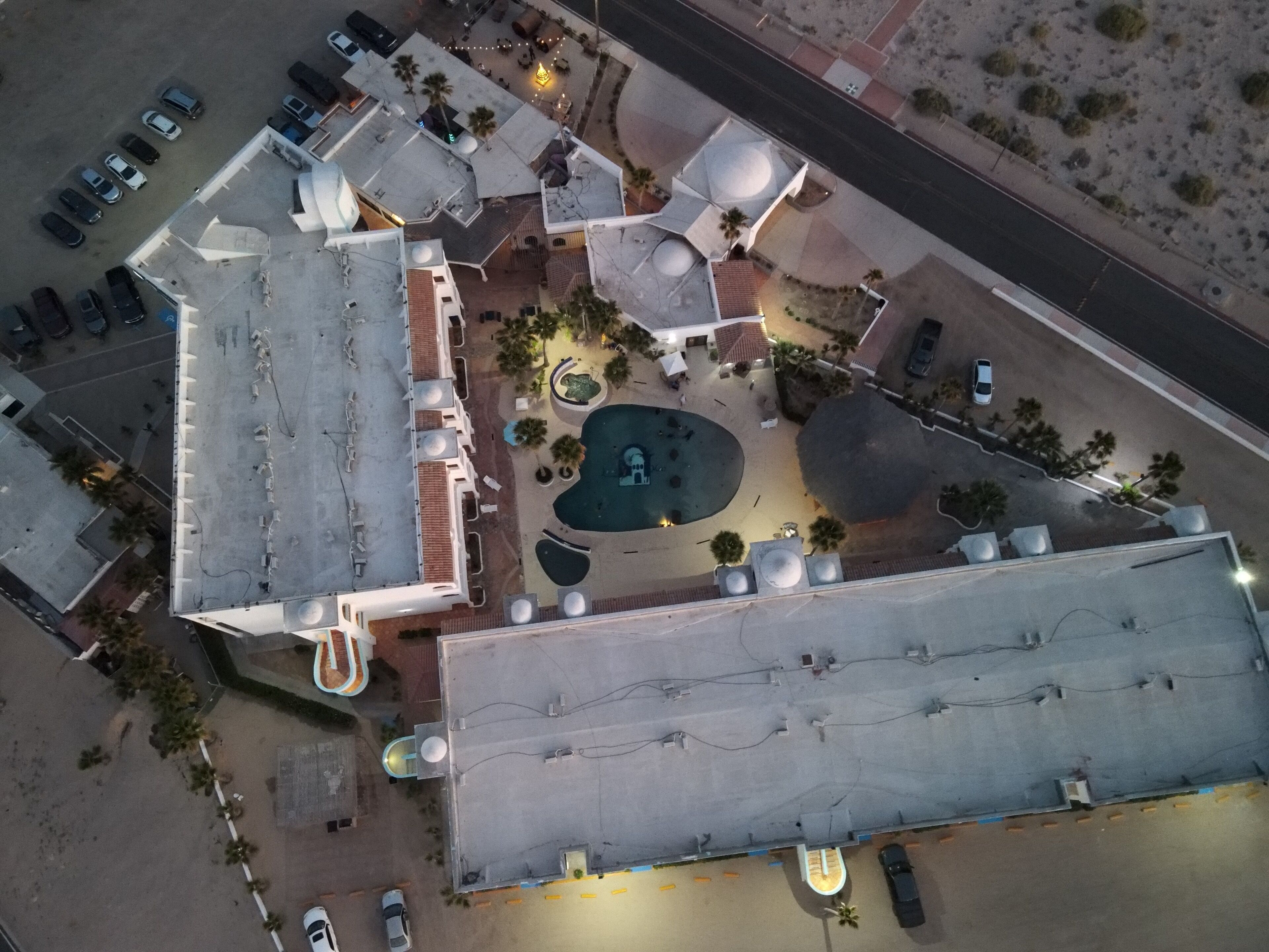 An aerial view of a hotel with a rooftop pool, multiple parking lots, and a central courtyard with palm trees.