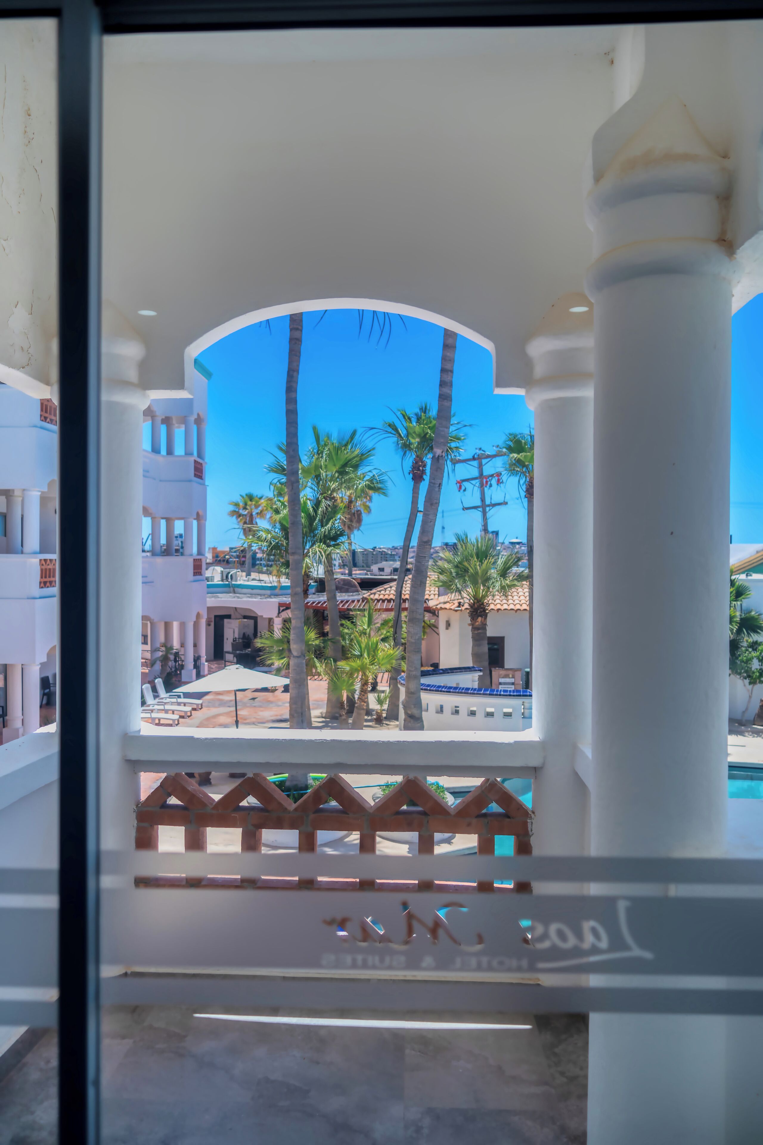 A view from a balcony with a white railing, palm trees, and a building with a blue roof in the background.