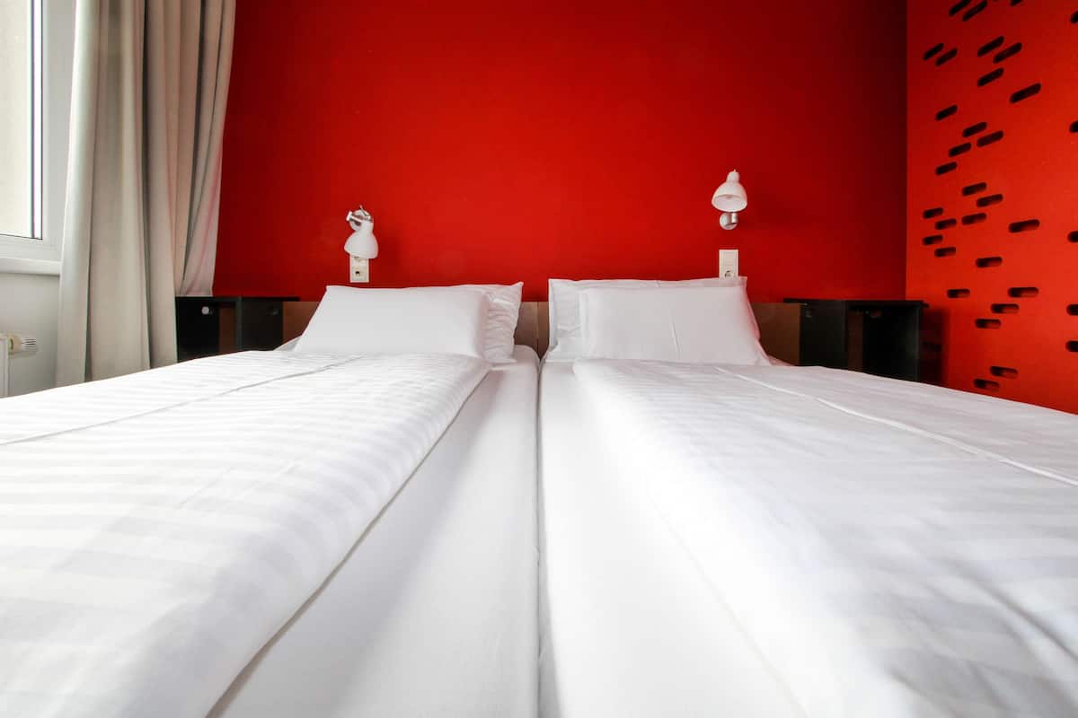 A double bed in a room with a red wall and a window with curtains.