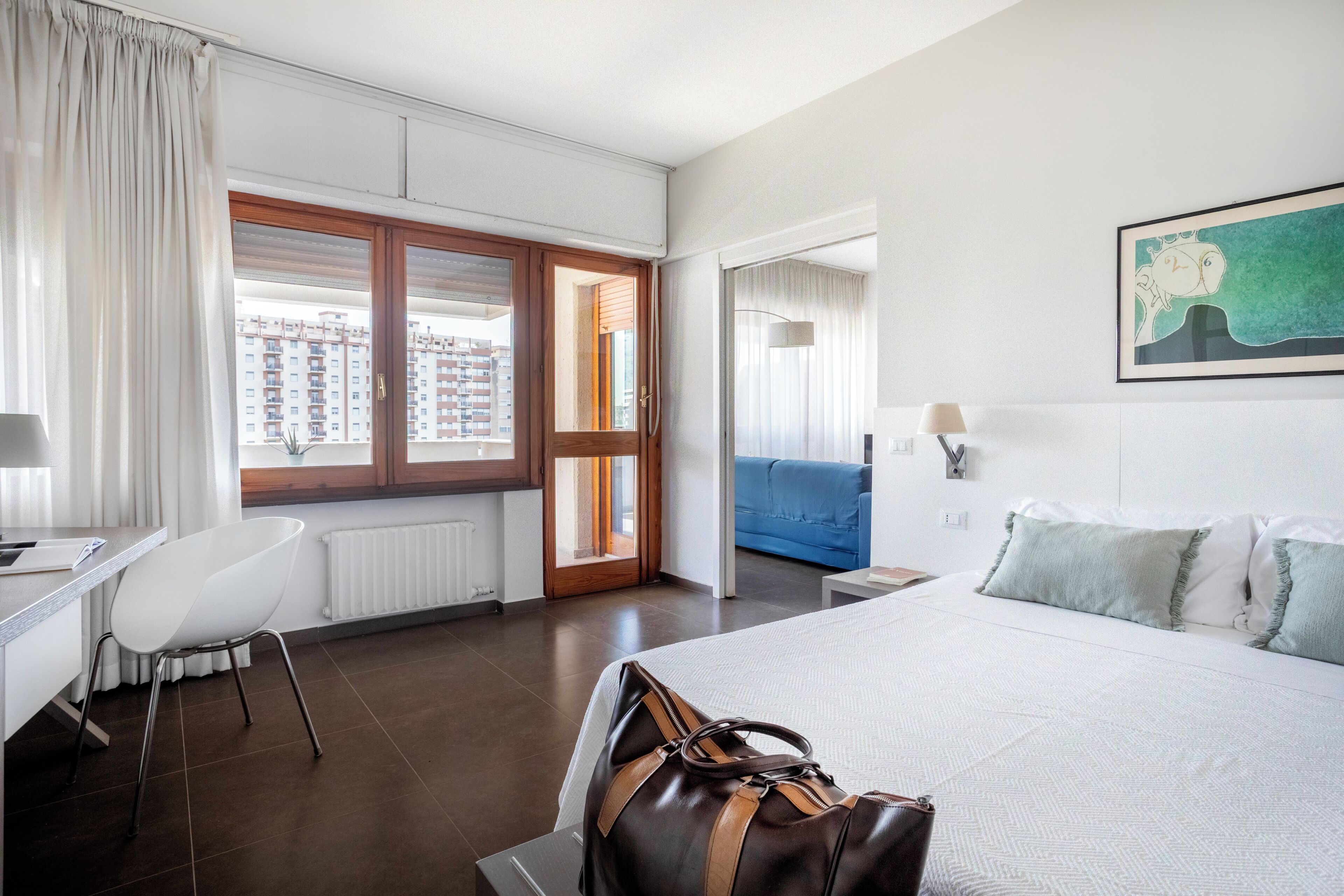 A hotel room with a bed, a desk, a chair, a window with curtains, and a painting on the wall.