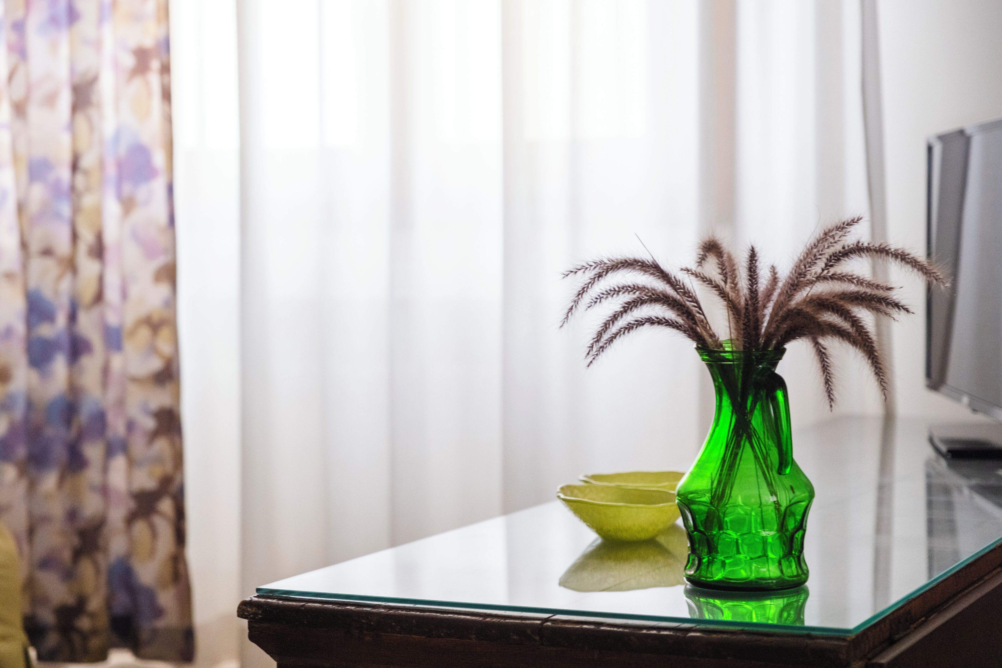 Un florero verde con plantas secas sobre una mesa de vidrio reflectante.