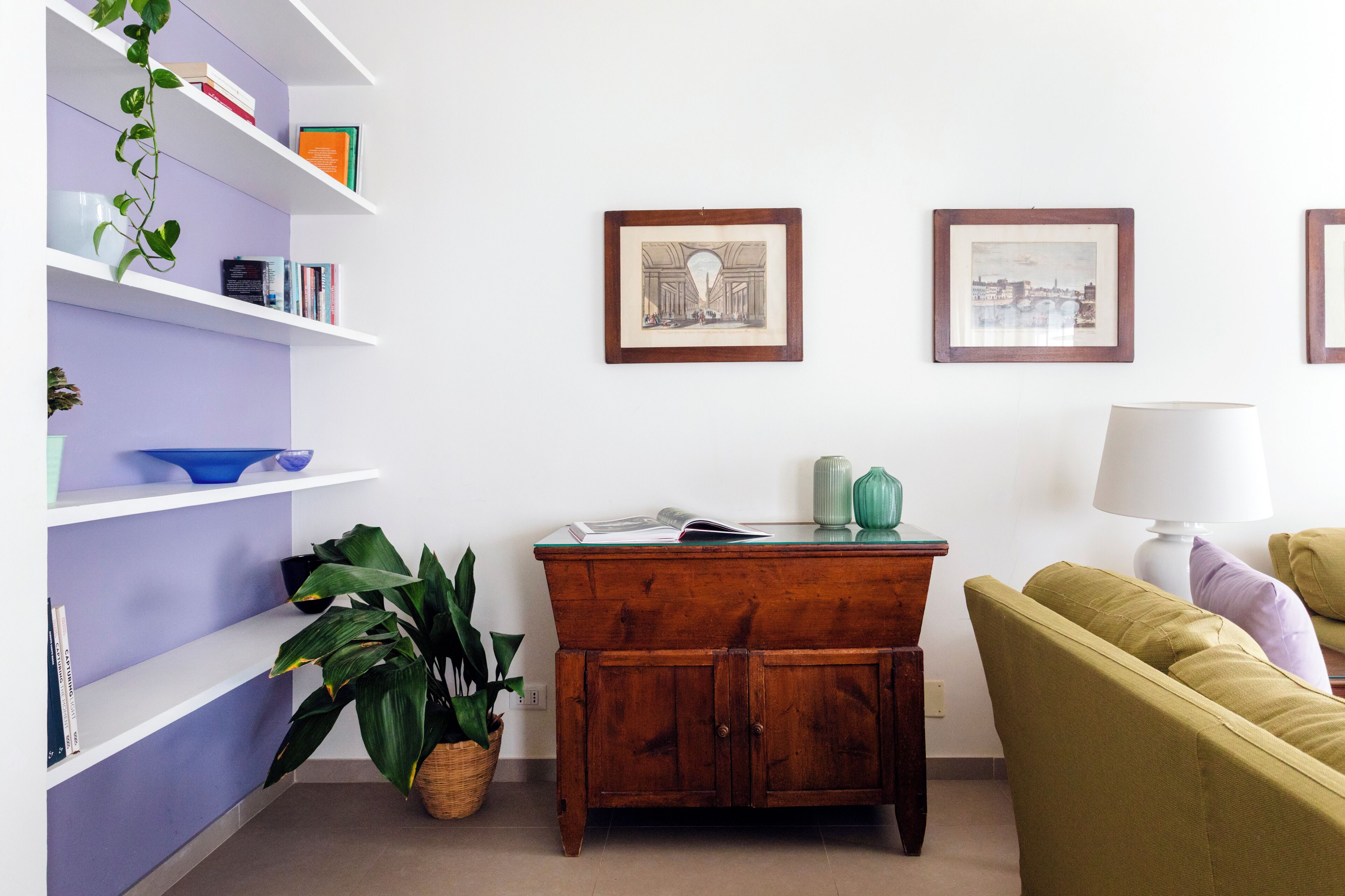 A room with a wooden cabinet, a green sofa, a white lamp, and shelves with books and decorative items.