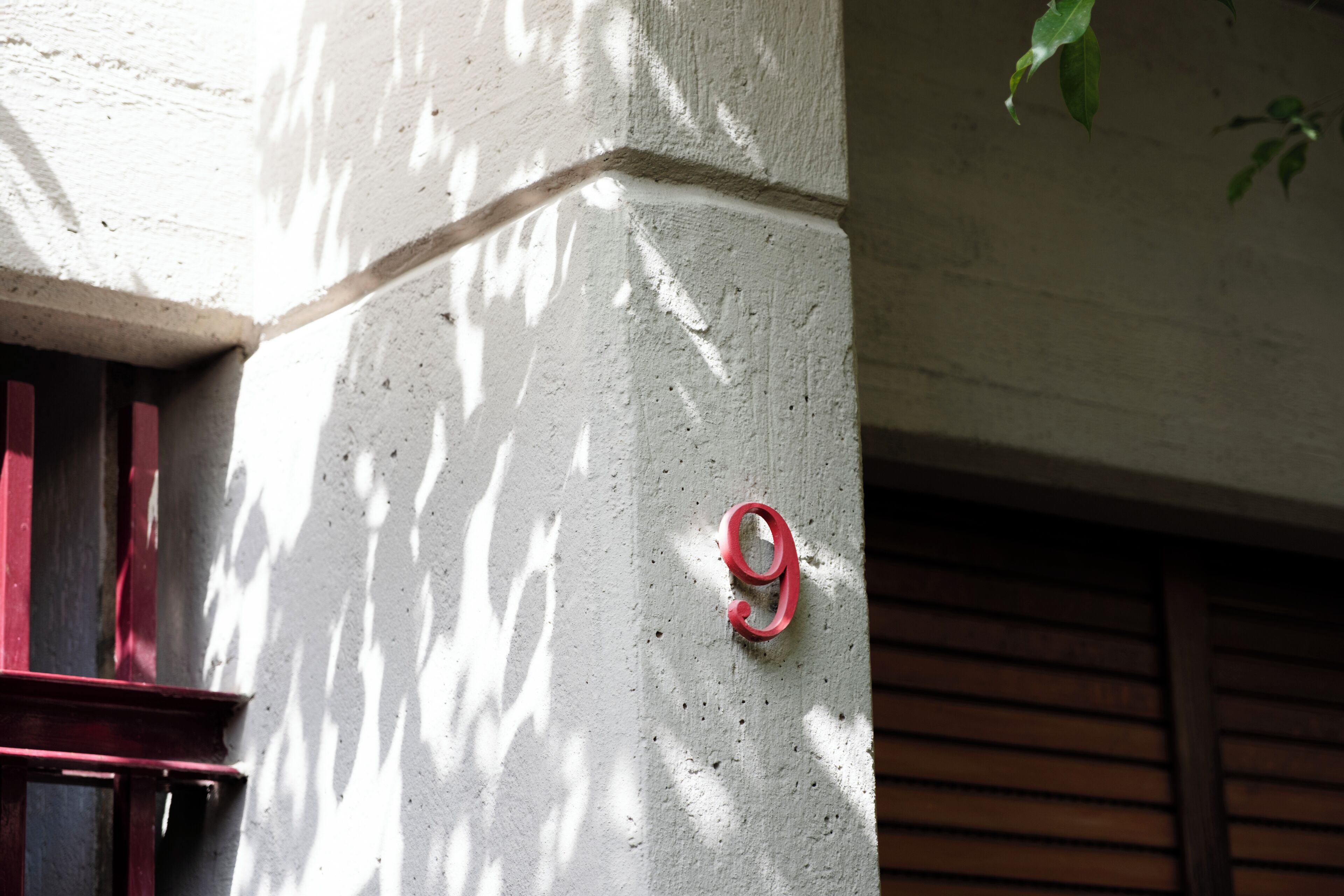 Un coin d’immeuble avec le chiffre « 9 » peint sur un mur, une fenêtre aux encadrements rouges et l’ombre d’un arbre.