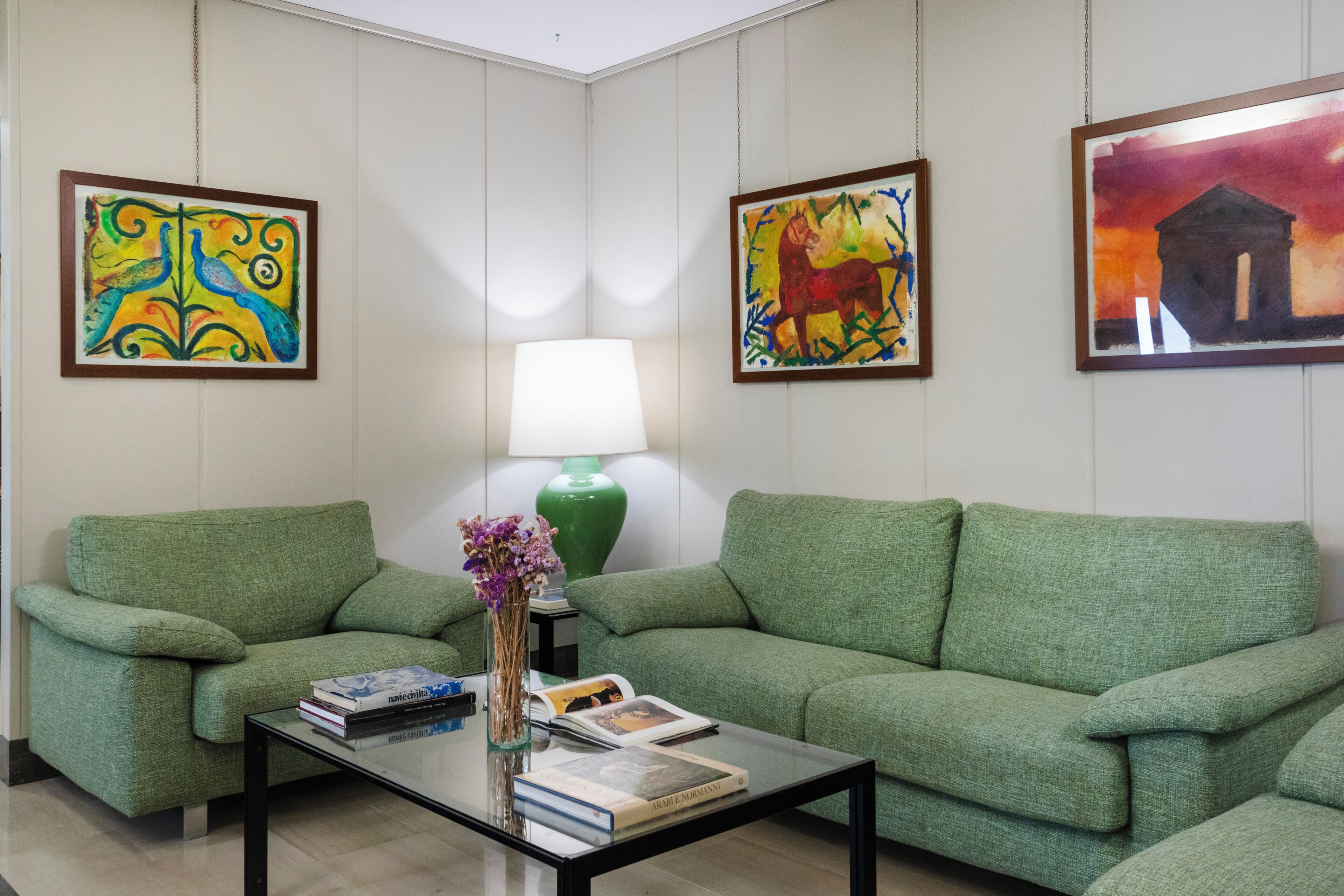 Una habitación con dos sofás verdes, una mesa de centro de vidrio con libros y un jarrón, una lámpara verde y tres obras de arte enmarcadas en las paredes.