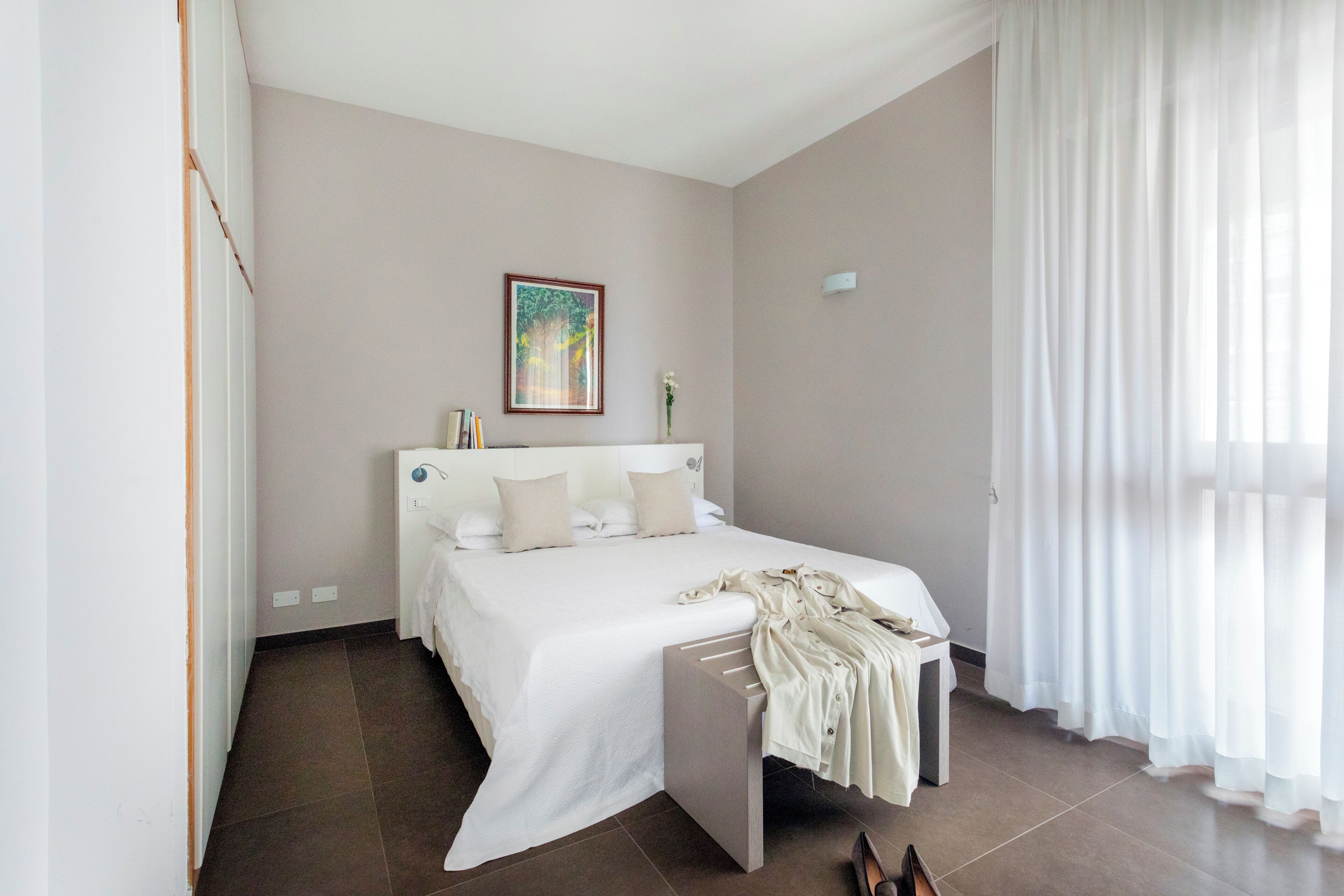 A bedroom with a bed, a bedside table, a nightstand, a painting on the wall, and a window with curtains.