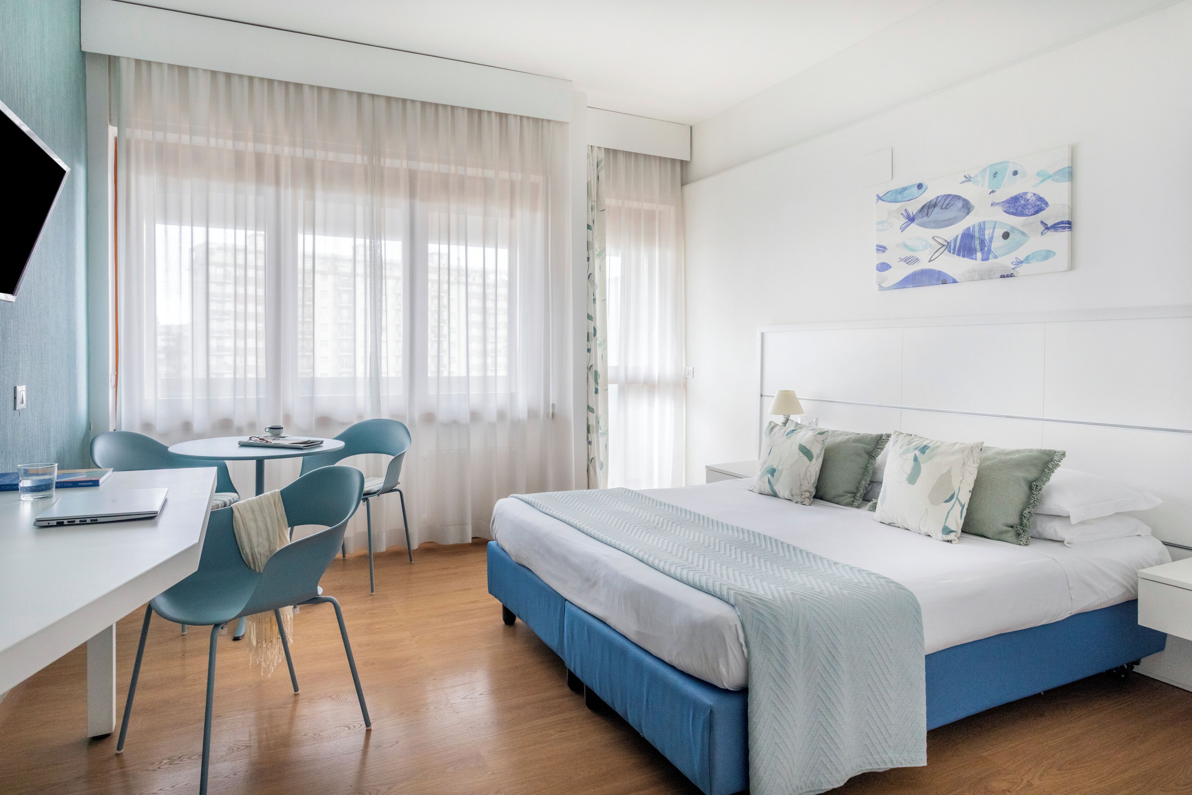 A hotel room with a bed, a desk, chairs, and a painting on the wall.
