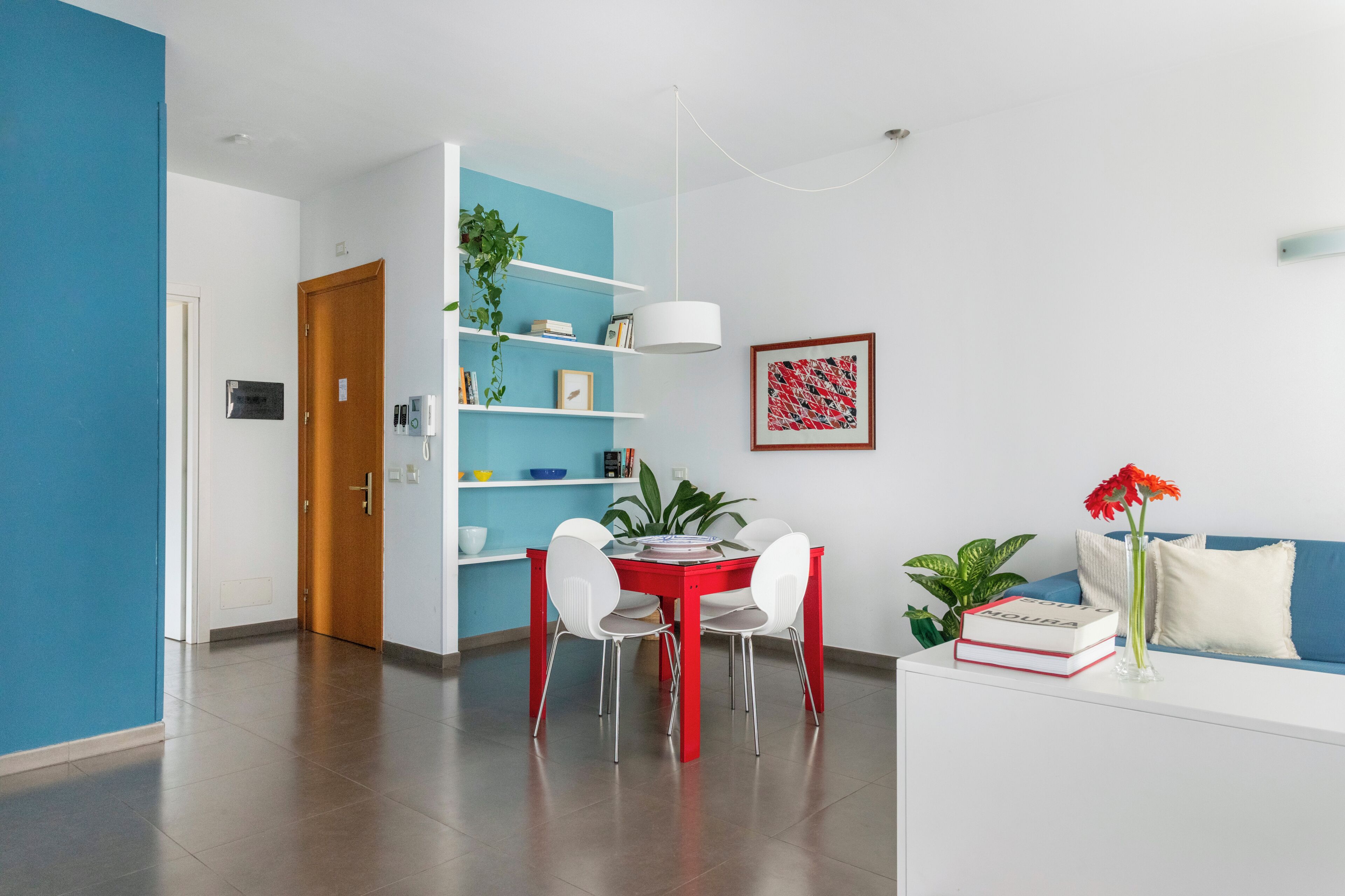 Un comedor moderno con una mesa roja y sillas blancas, una pared azul y un sofá blanco con un jarrón de flores.