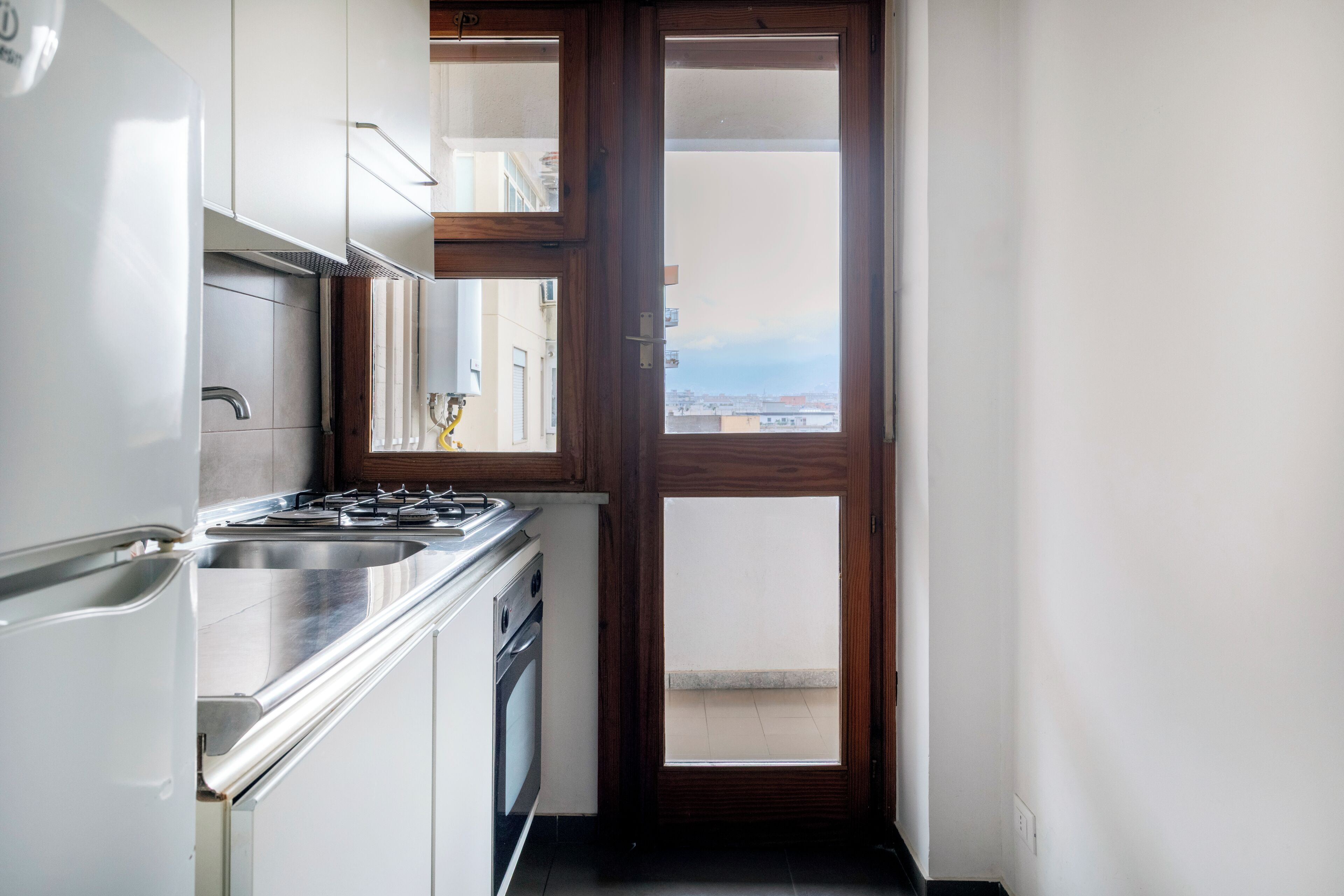 A narrow kitchen with a stove, sink, and cabinets, leading to a balcony with a city view.