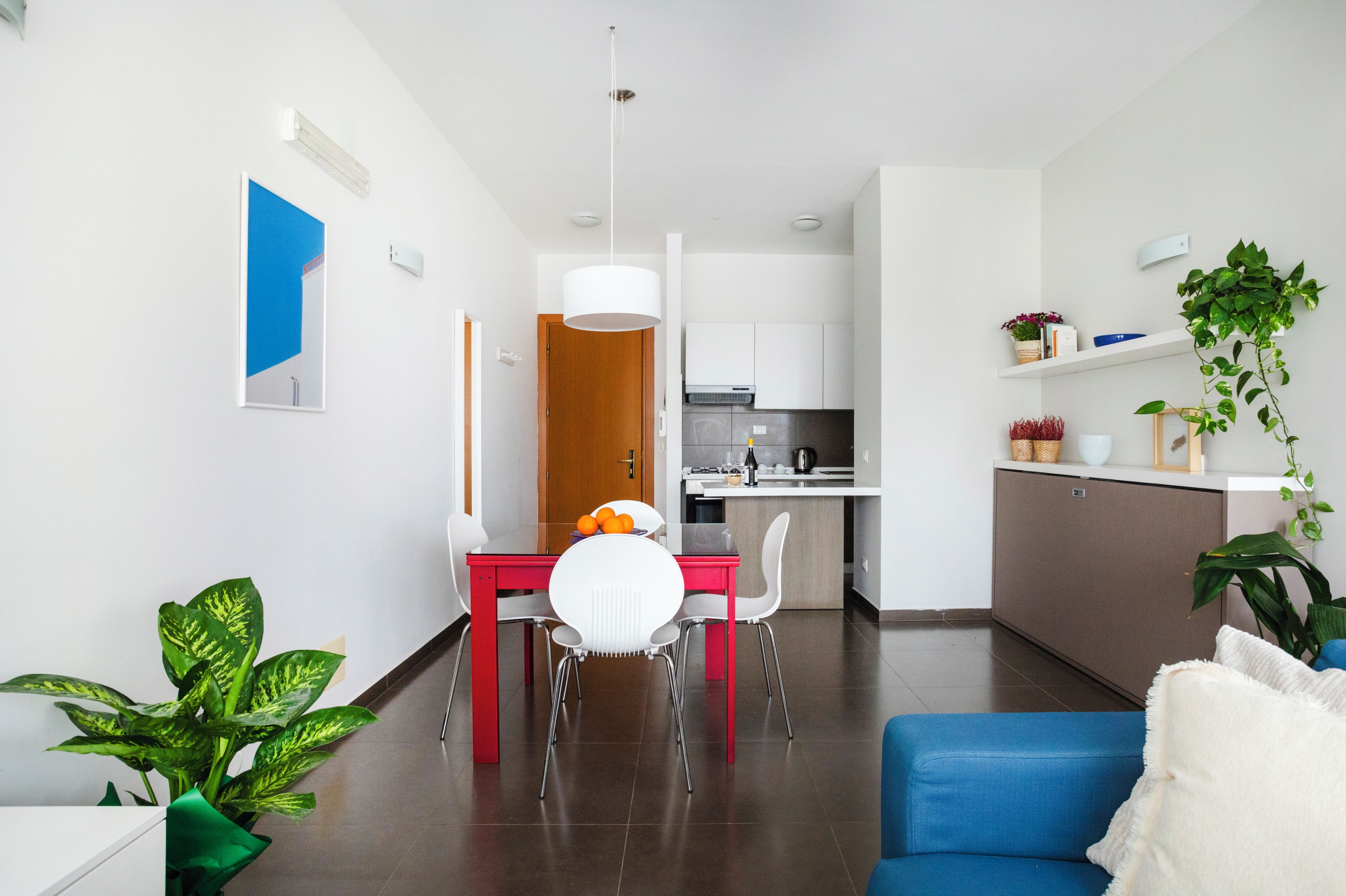 Un saló modern amb un sofà blau, una taula de dinar amb cadires, una cuina al fons i una planta en test.