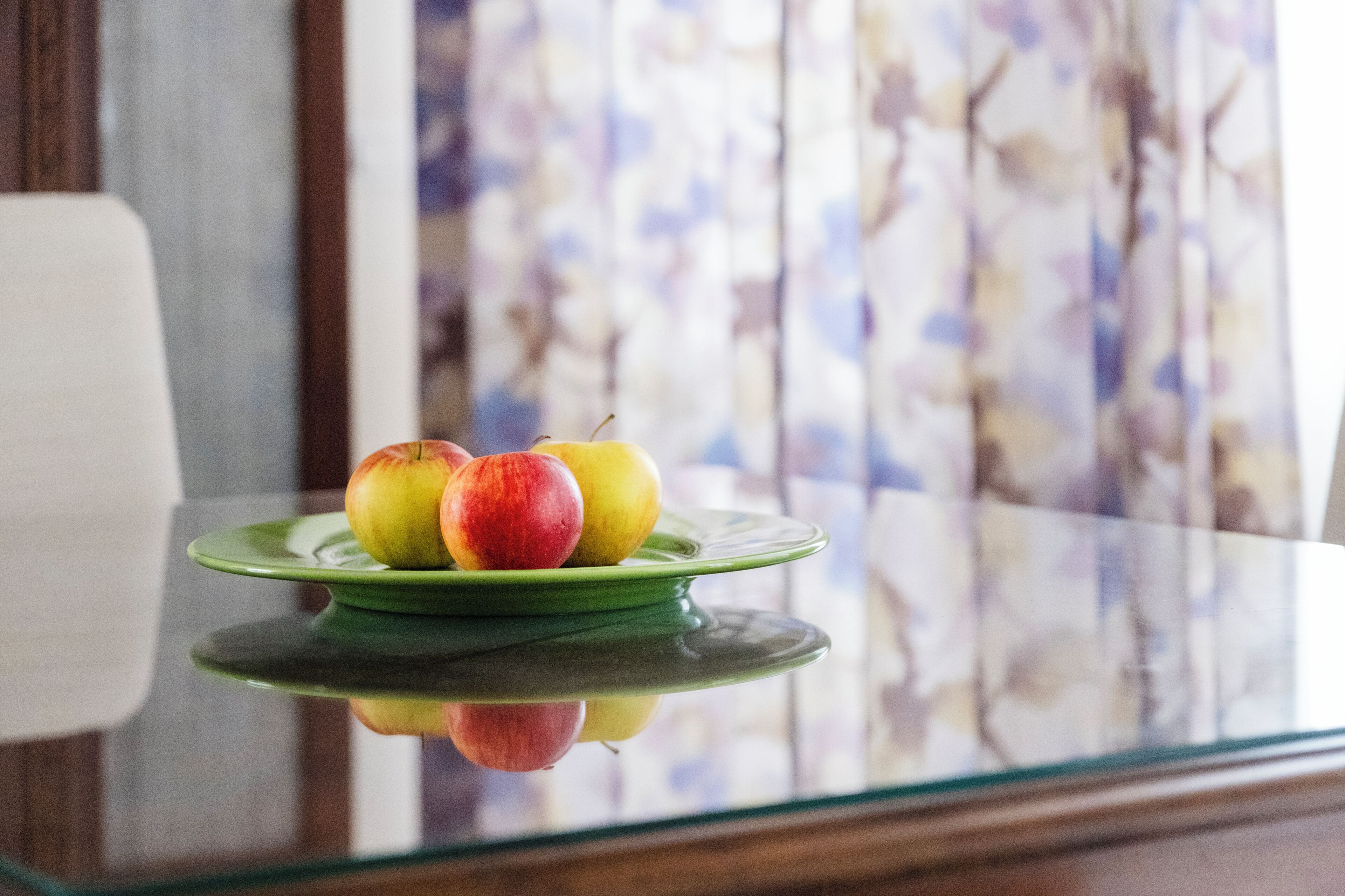 Una mesa de vidrio con un plato verde que sostiene tres manzanas.