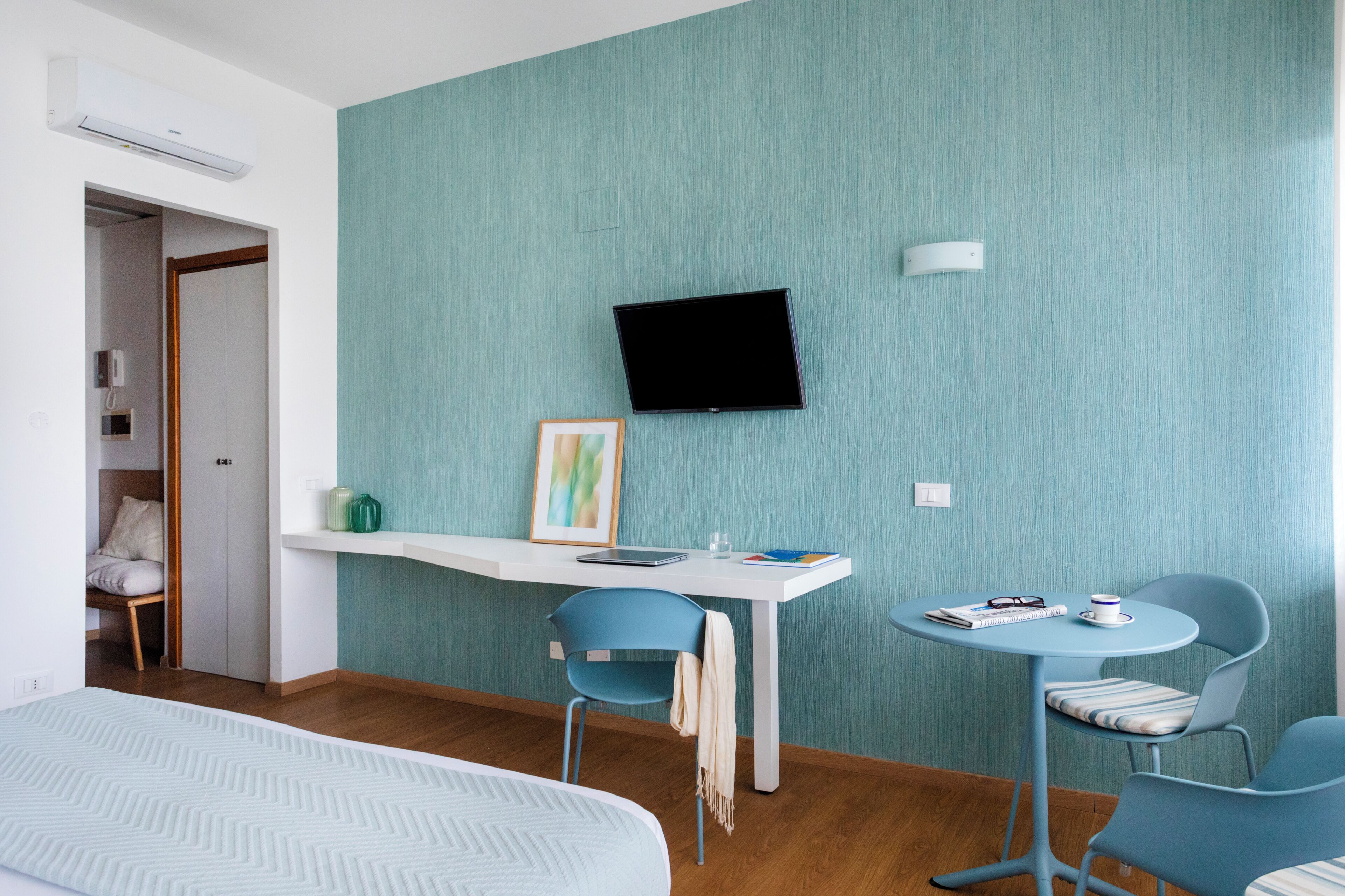 A modern hotel room with a bed, a desk with a chair, a TV mounted on the wall, and a small table with a chair.