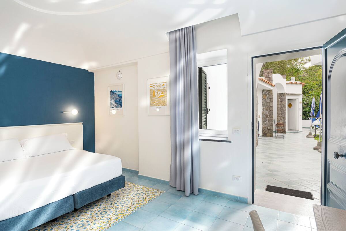 Un dormitorio con cama, ventana con cortinas, puerta que da a un área exterior y pared con cuadros.
