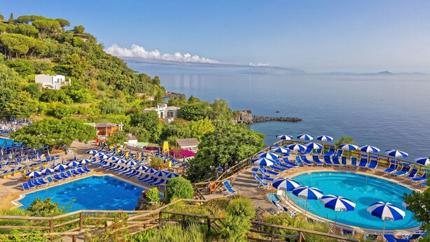 Un complexe hôtelier doté de plusieurs piscines, de chaises longues et de parasols, entouré d’une végétation luxuriante et offrant une vue sur une mer calme.