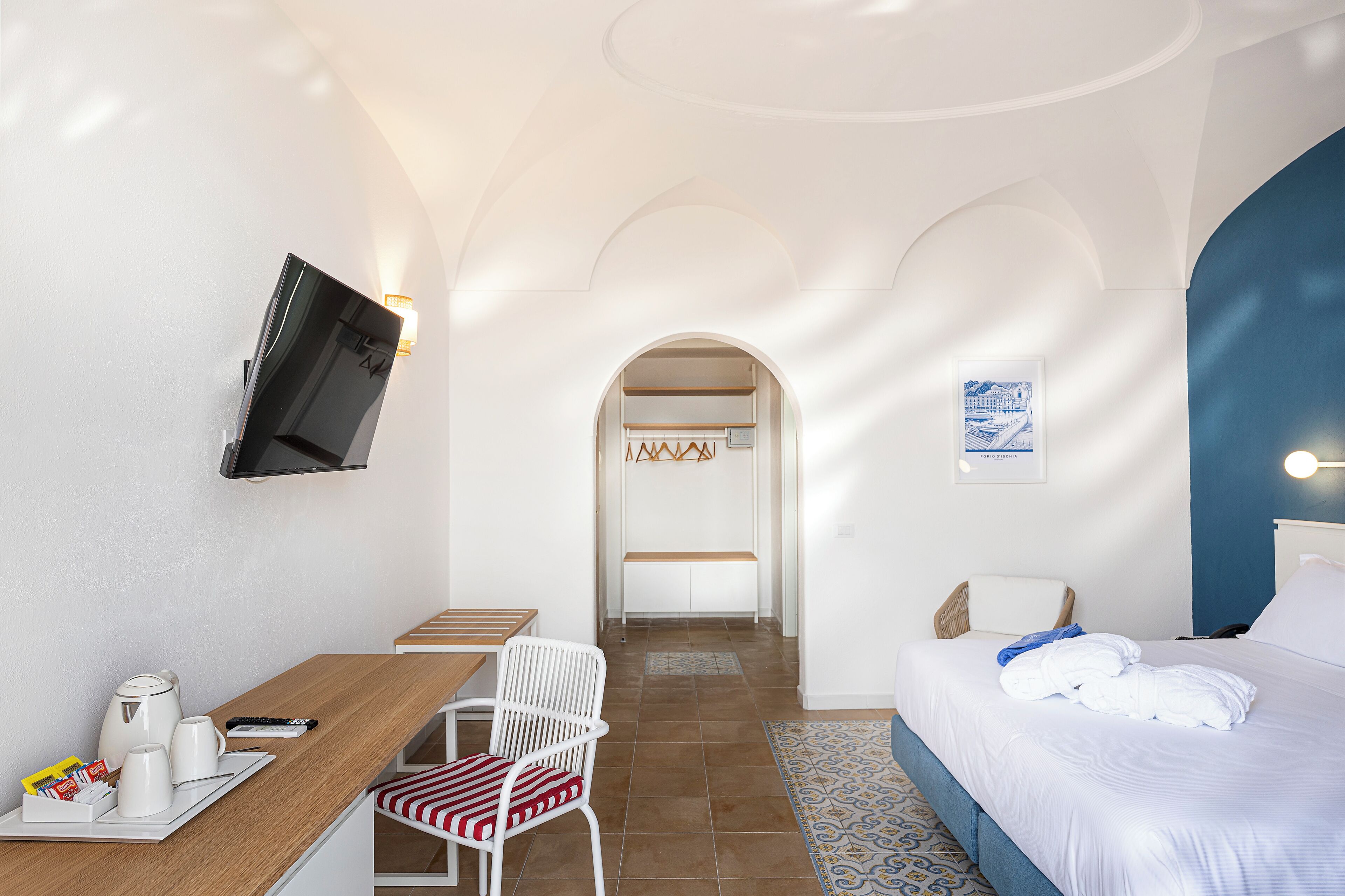 A hotel room with a bed, a desk with a chair, a TV mounted on the wall, and a bathroom visible through an archway.