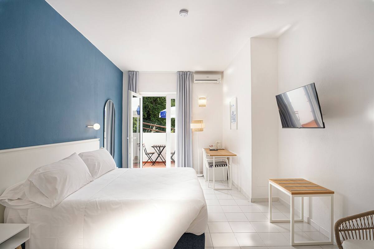 Habitación de hotel con cama, escritorio, silla, televisor y balcón con vistas a zonas verdes.