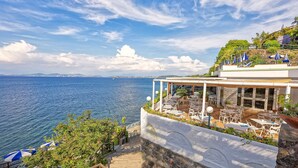 Une terrasse en bord de mer avec des places assises à l’extérieur, une vue sur la mer et une côte visible au loin.