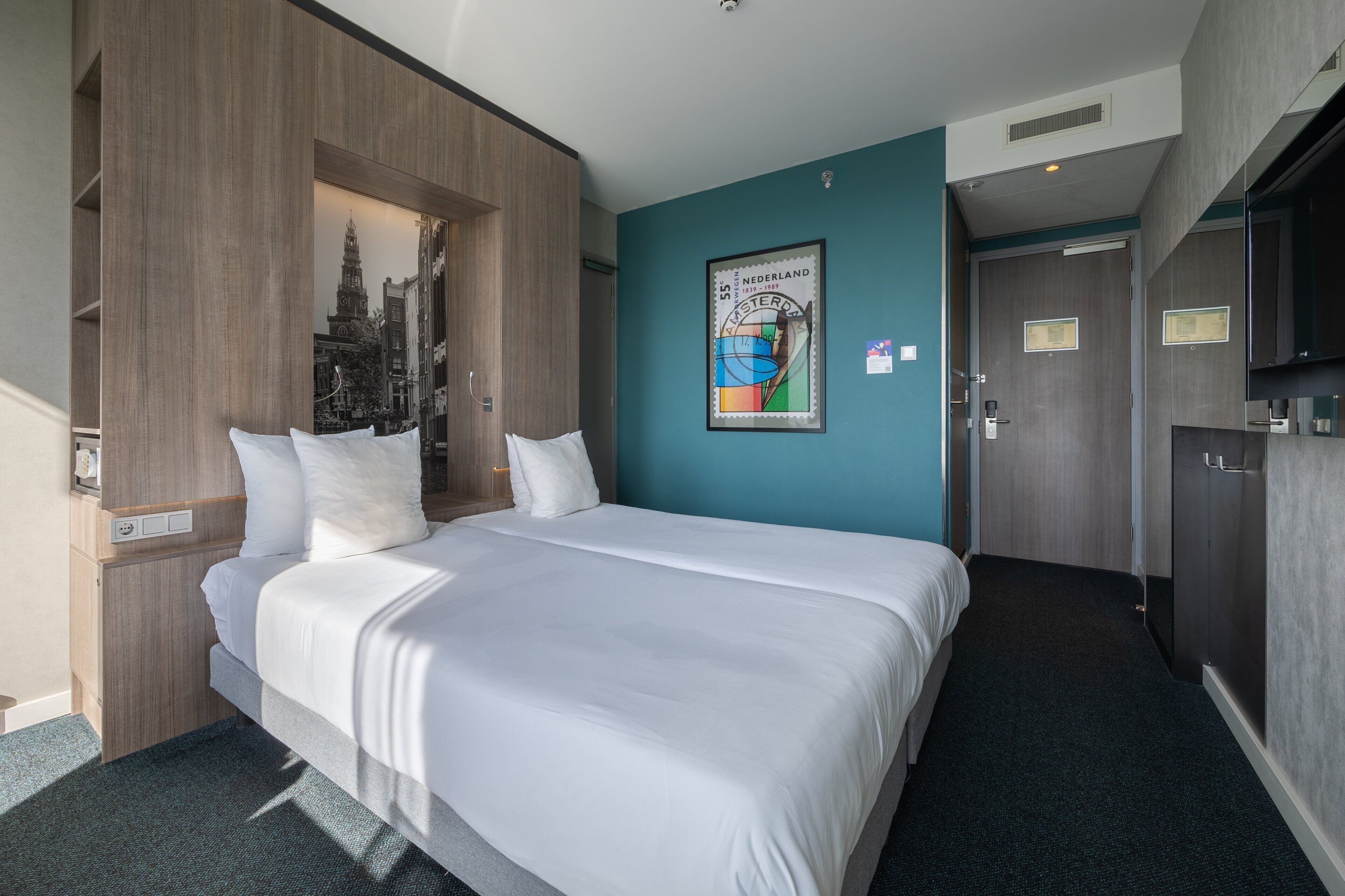 Quarto de hotel com uma cama grande, um guarda-roupa de madeira, uma parede de destaque azul-esverdeada e um cartaz emoldurado com vista para a cidade.