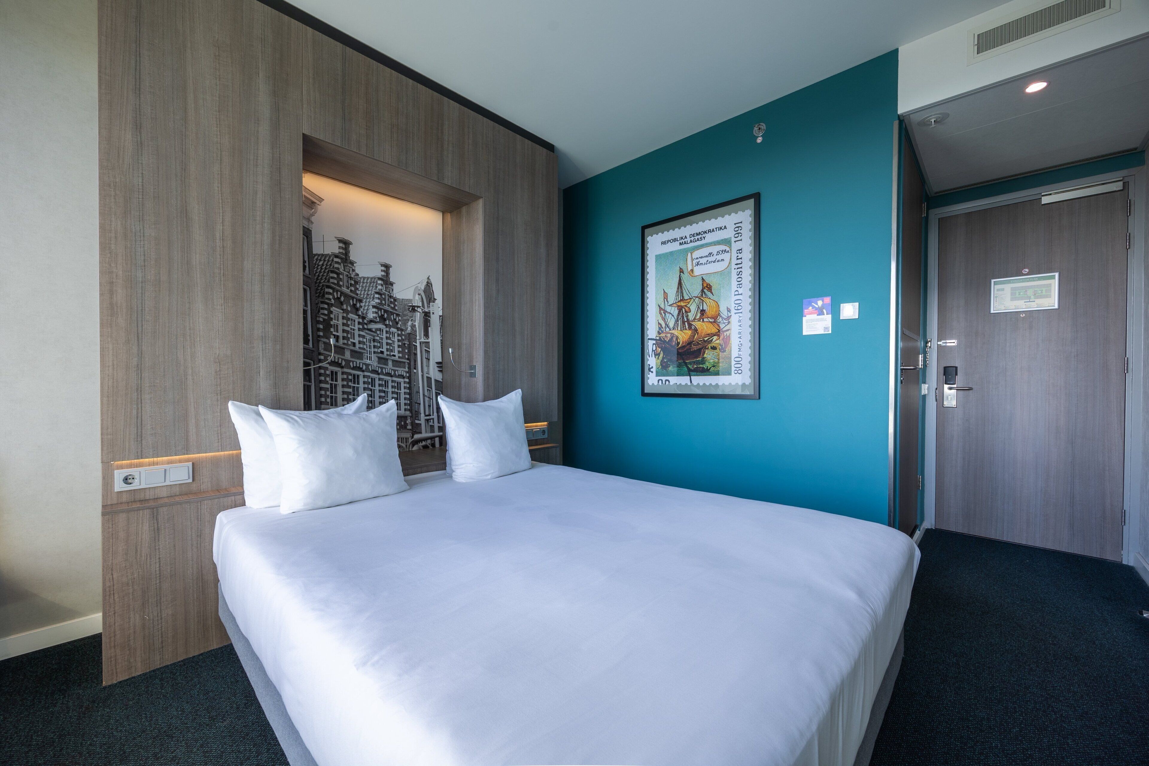 Ein Hotelzimmer mit einem großen Bett, einem Stadtbild-Kunstwerk, einer gerahmten Karte und einer Tür mit einem Schild.