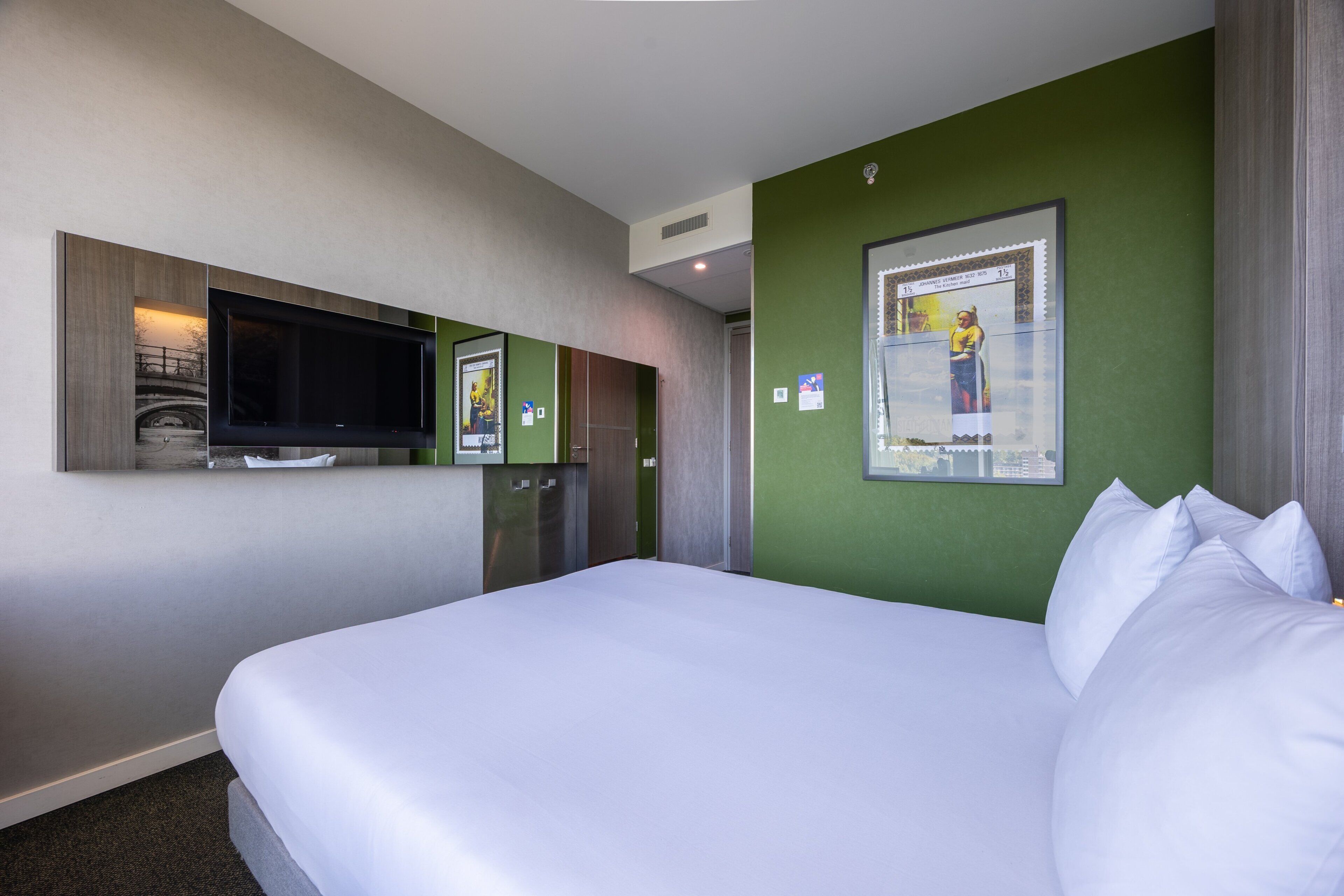 Habitación de hotel con una cama grande, un televisor de pantalla plana montado en la pared y un cuadro enmarcado sobre la cama.