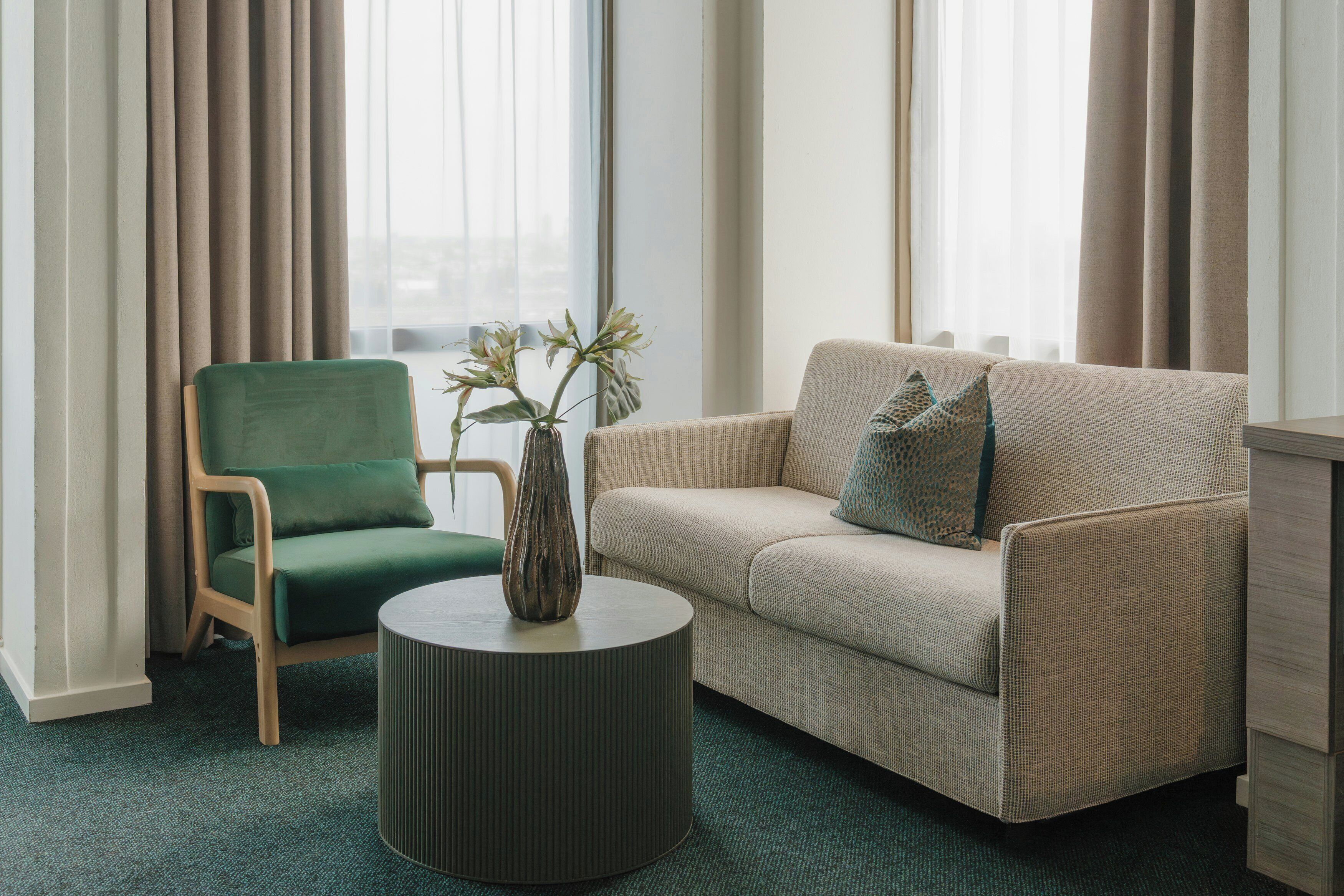 A room with a green chair, beige sofa, and a round table with a vase.