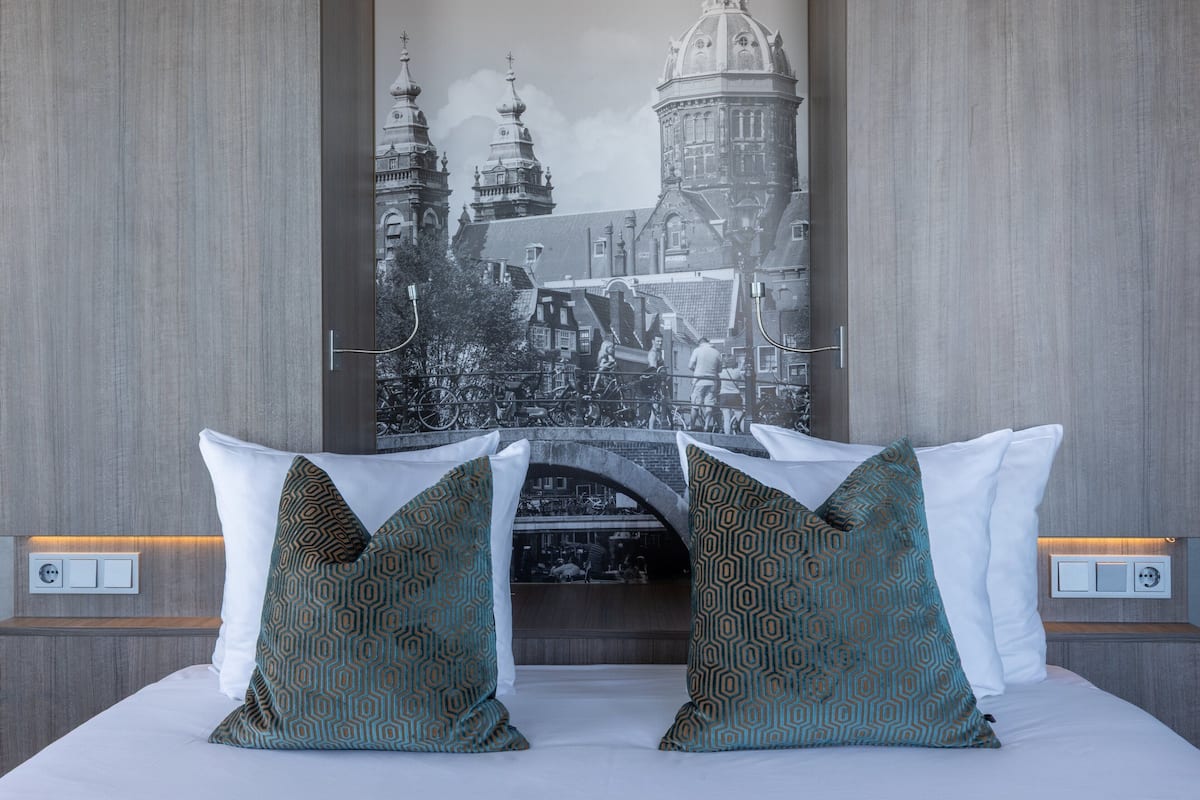 Ein Hotelzimmer mit einem großen Bett, zwei gemusterten Kissen, einem wandmontierten Lichtschalter und einem gerahmten Bild eines historischen Gebäudes.