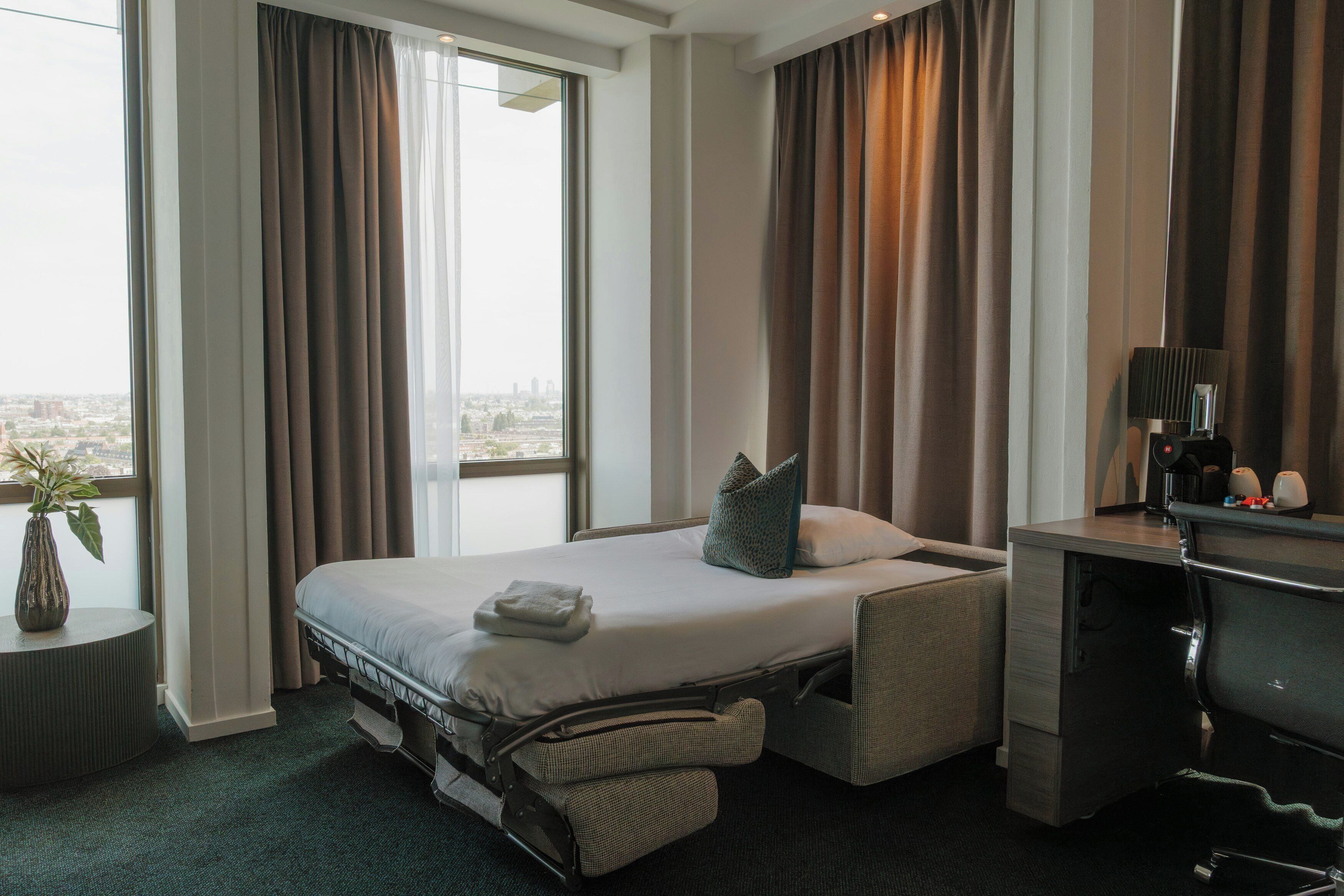 A hotel room with a bed, a desk, a chair, a lamp, a plant, and a view of the city.