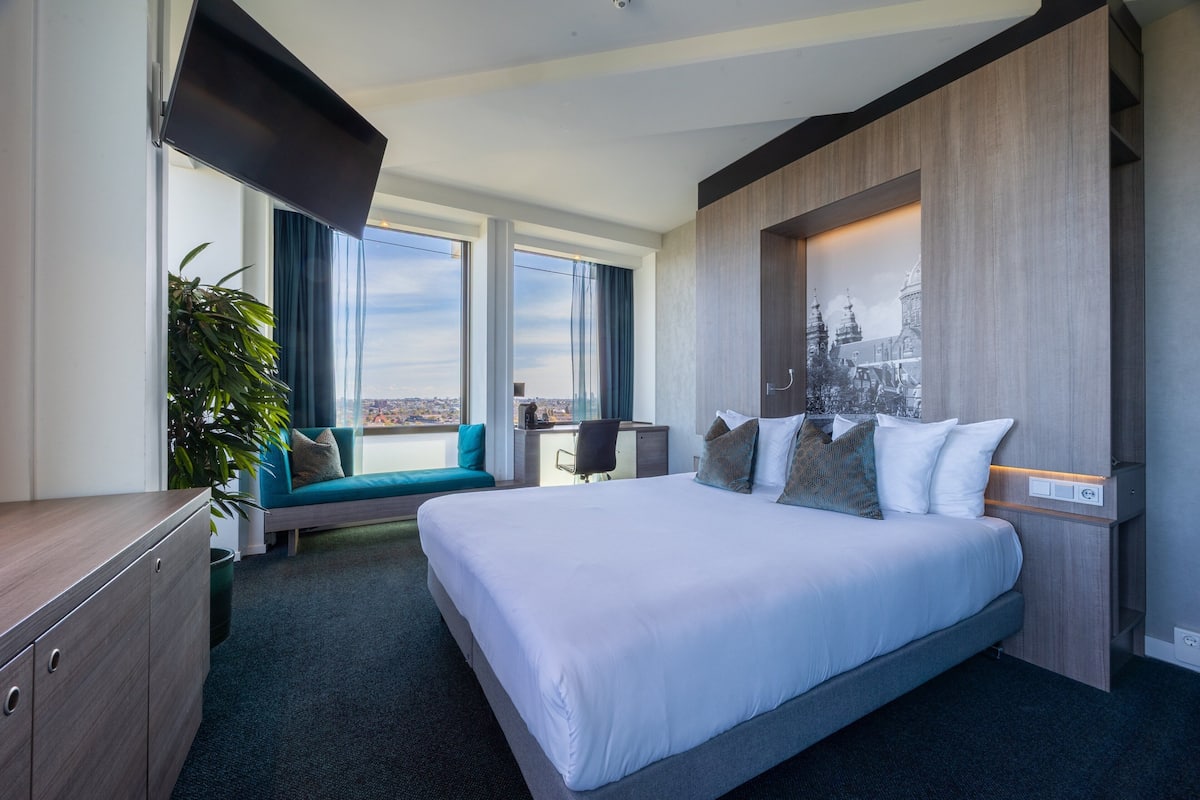 A modern hotel room with a large bed, a desk, a seating area with a view, and a wall-mounted TV.