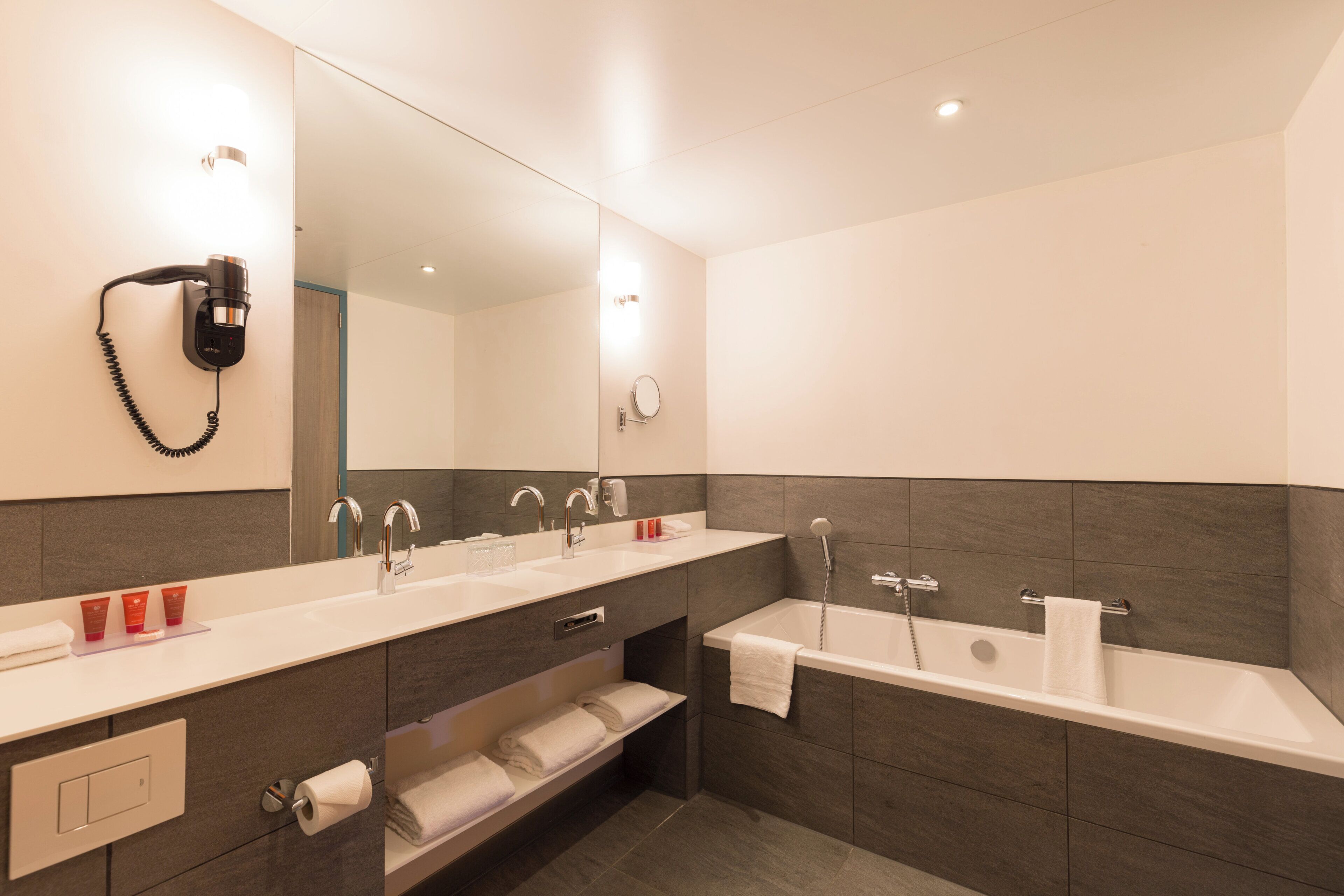 A modern bathroom with a double sink, a bathtub, a hairdryer mounted on the wall, and a large mirror.