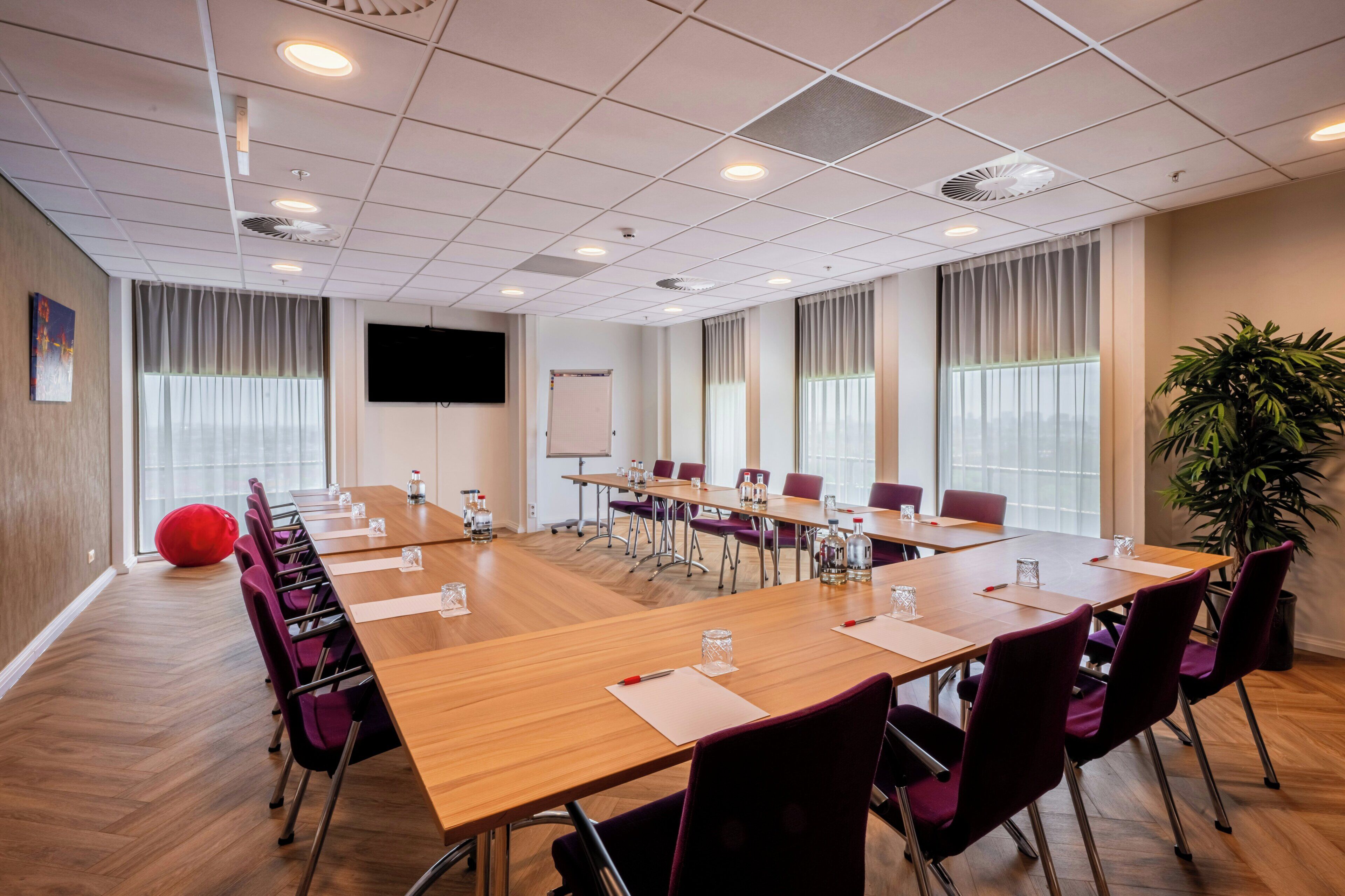 Un salón de conferencias con una larga mesa, sillas, una pantalla de proyector y una planta grande.