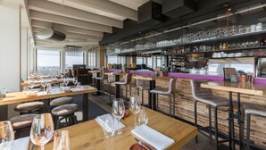 Un restaurante moderno con mesas de madera y asientos en la barra, un expositor de vidrio con copas y una zona de bar con botellas y copas.