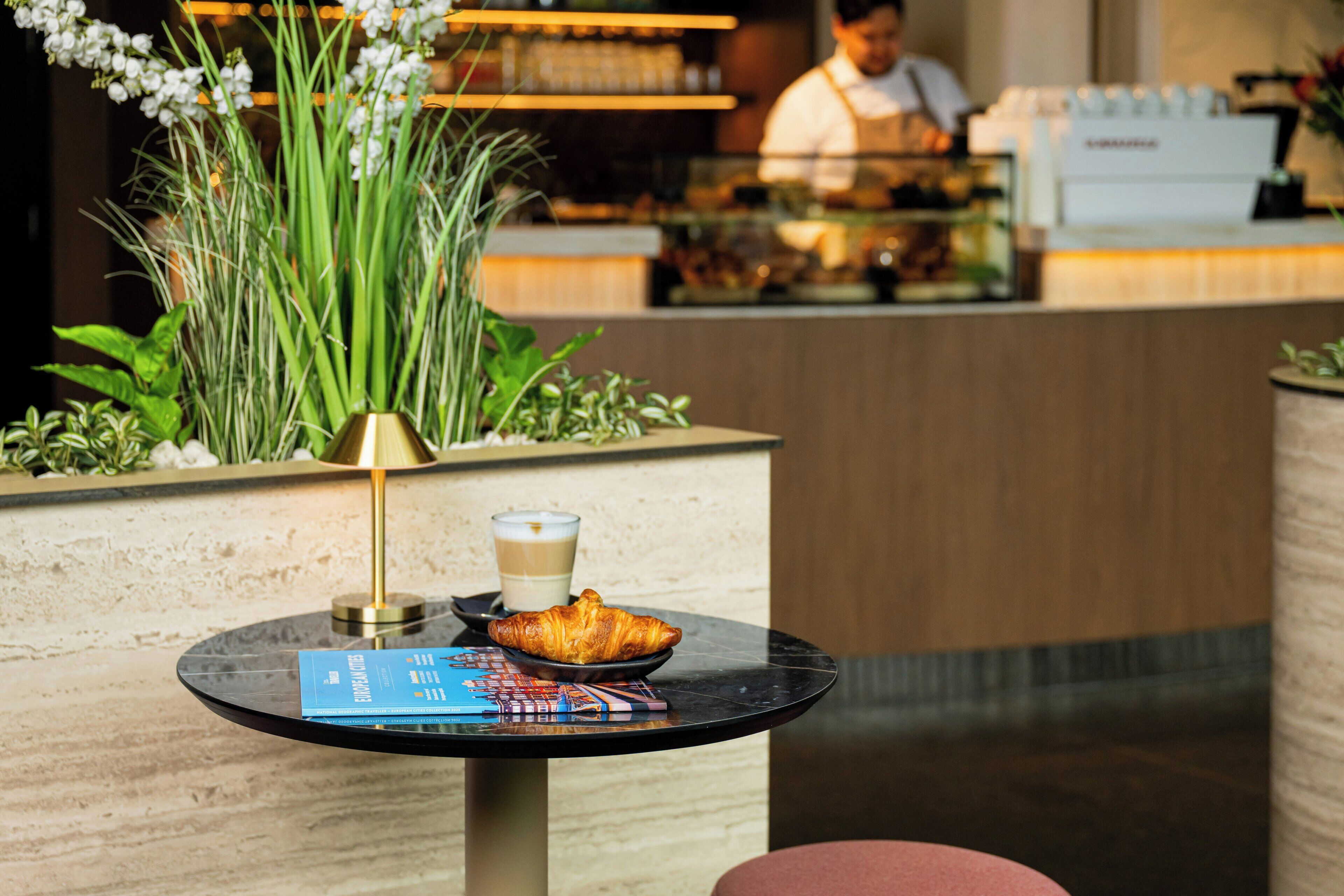 A table with a croissant, a cup of coffee, and a book in a modern setting with a bar and a chef in the background.