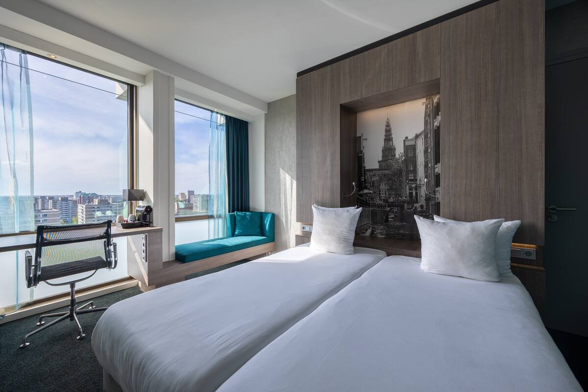 Ein Hotelzimmer mit einem großen Bett, einem Schreibtisch mit Stuhl, einer Sitzgelegenheit am Fenster und Blick auf die Stadt.