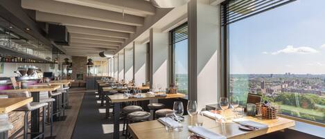 Un restaurante moderno con mesas largas y asientos de bar, grandes ventanales que ofrecen vistas a la ciudad.