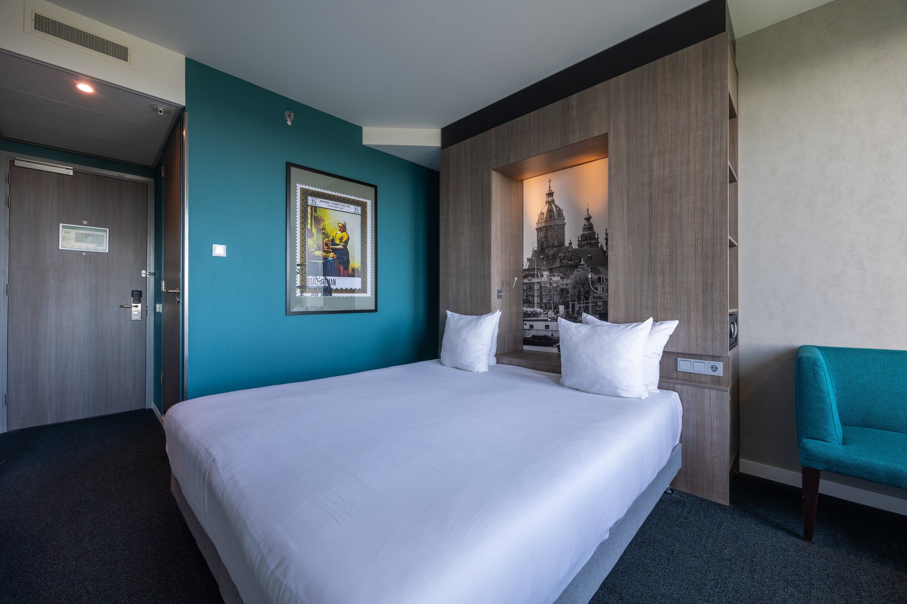 A hotel room with a large bed, a teal accent wall, a framed picture of a cityscape, and a teal armchair.